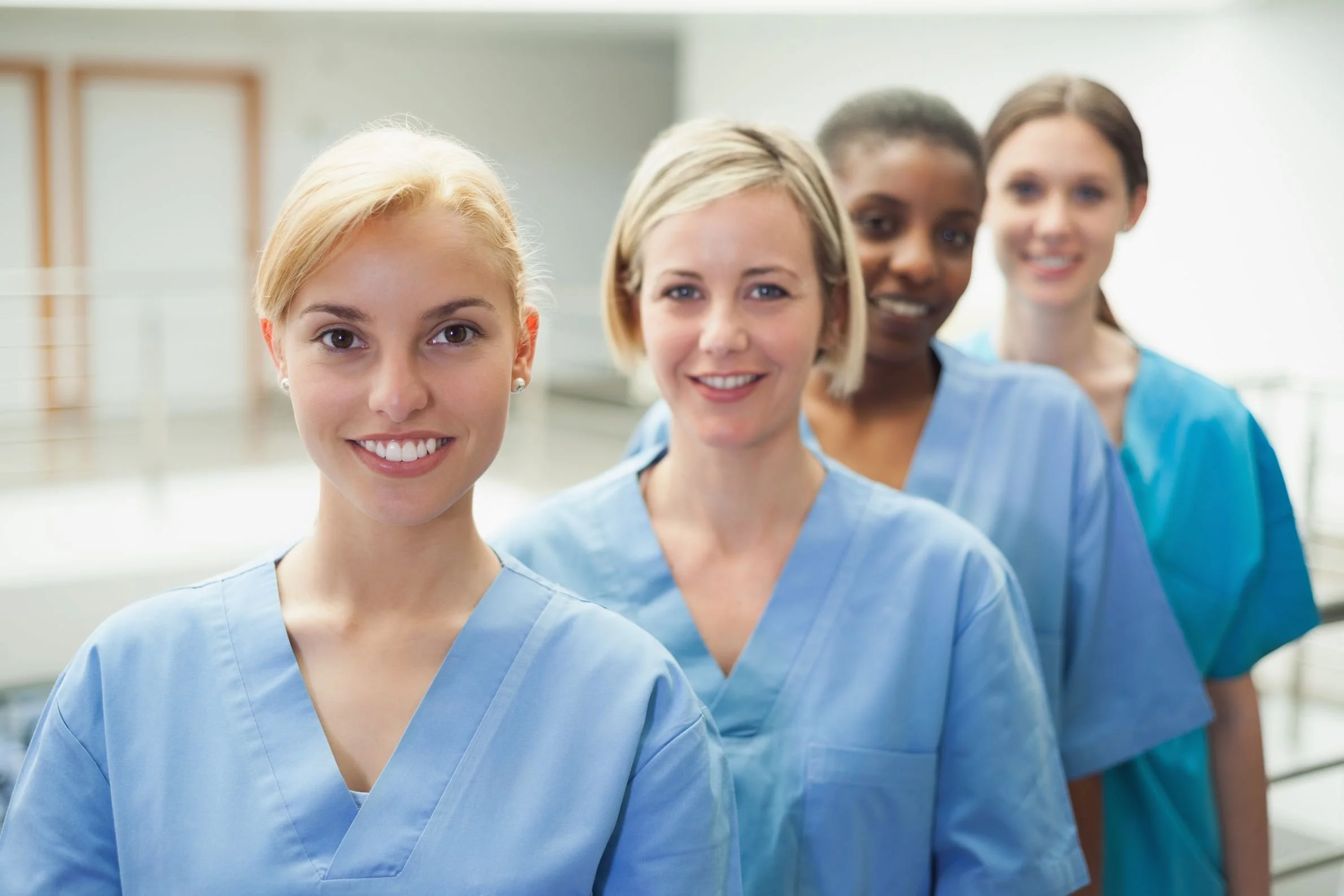 Four women in light blue scrubs standing in a line indoors, smiling at the camera.