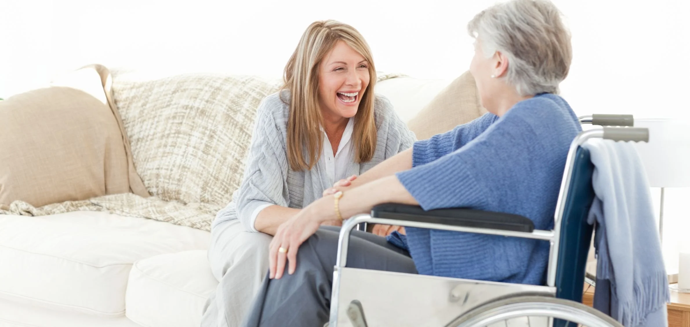 A woman with blonde hair laughing and holding hands with an elderly woman in a wheelchair, in a brightly lit living room.