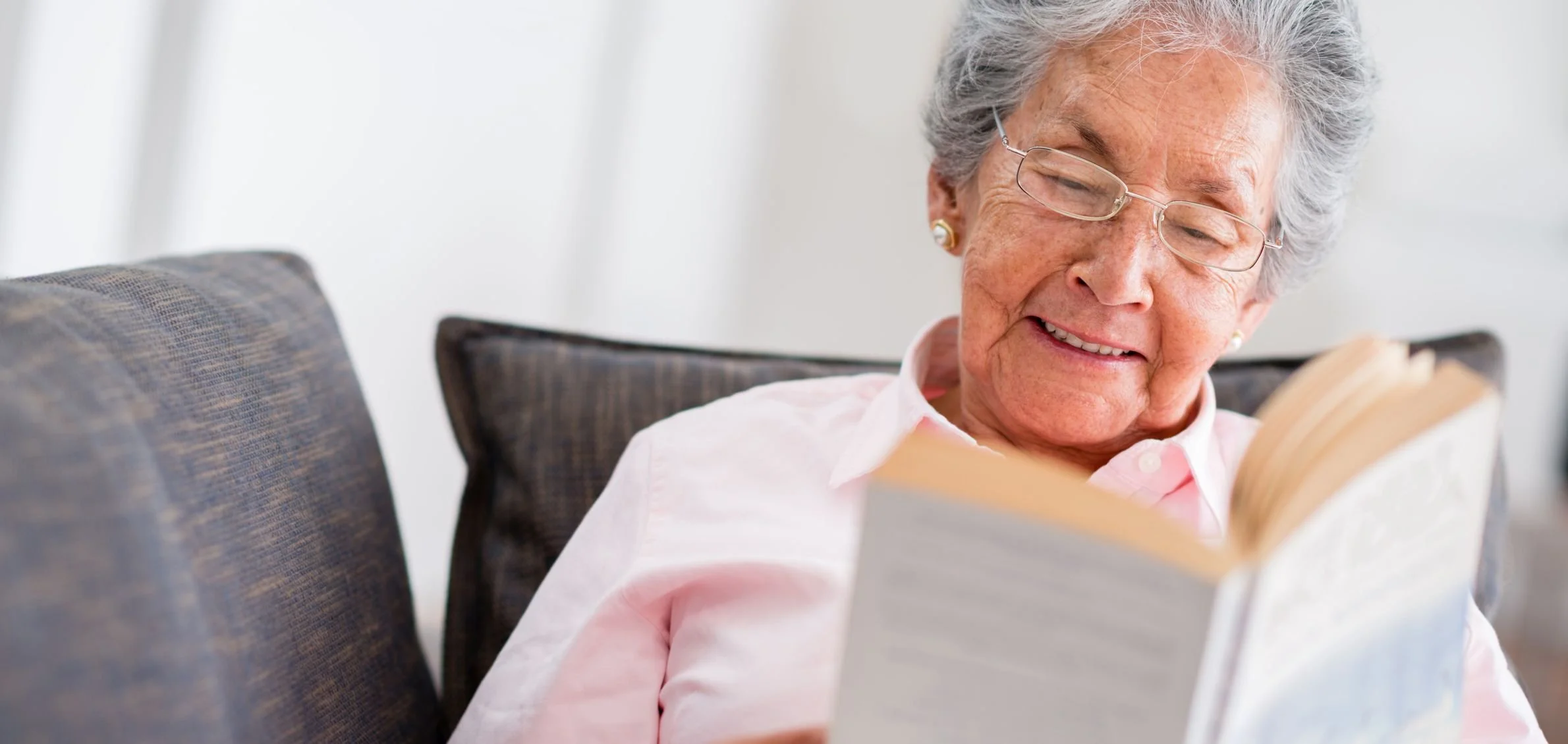 An elderly woman with gray hair, wearing glasses, reading a book while sitting on a sofa.