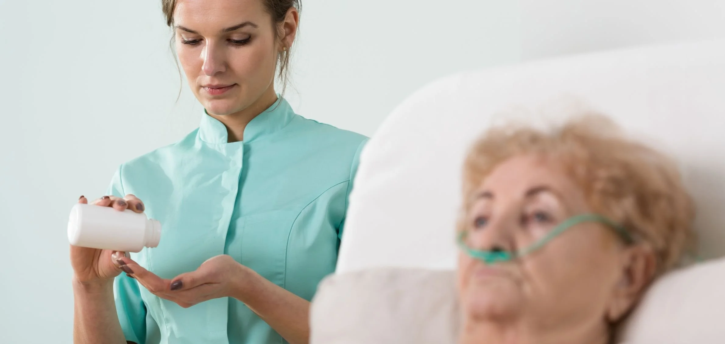 A middle-aged nurse in a teal uniform holding a pill bottle, looks down with a gentle expression. An elderly woman with light skin, curly hair, and wearing glasses lies in a hospital bed, partially visible, with her head on a pillow.