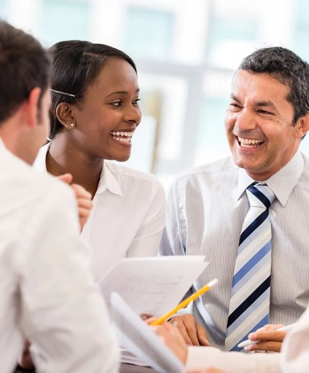 Three people in a professional meeting, smiling and engaging in conversation, with documents and a pencil on the table.