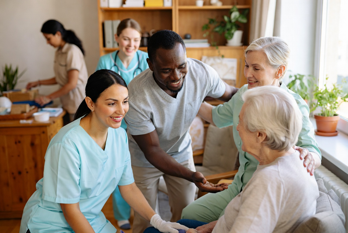 Group of healthcare workers, including nurses and a doctor, smiling and interacting with an elderly woman in a care home or hospital setting.