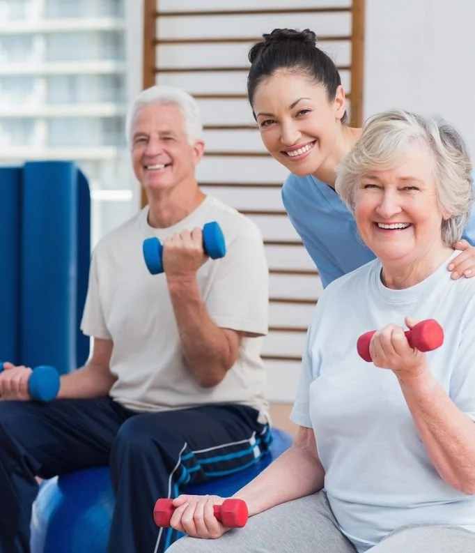 An elderly man and woman exercising with small dumbbells, guided by a young female trainer in a fitness center.