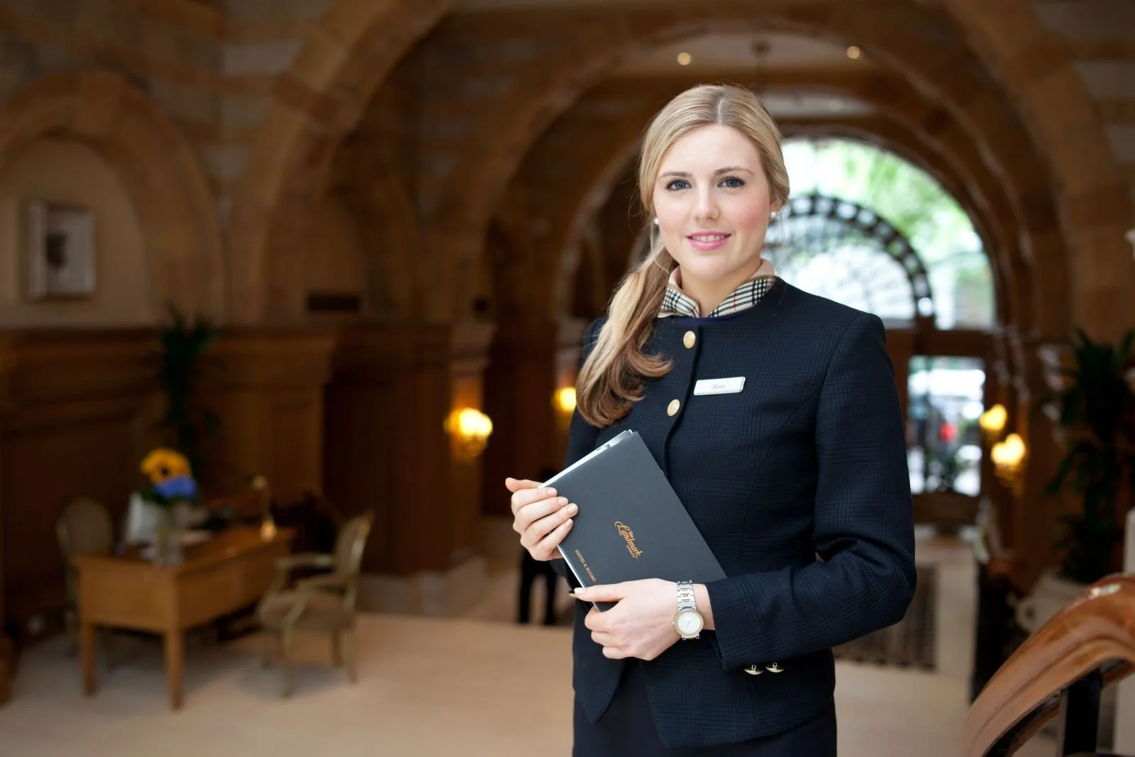 A smiling woman in a formal suit holding a folder inside a warmly lit, elegant restaurant or hotel lobby with wooden arches and furniture.