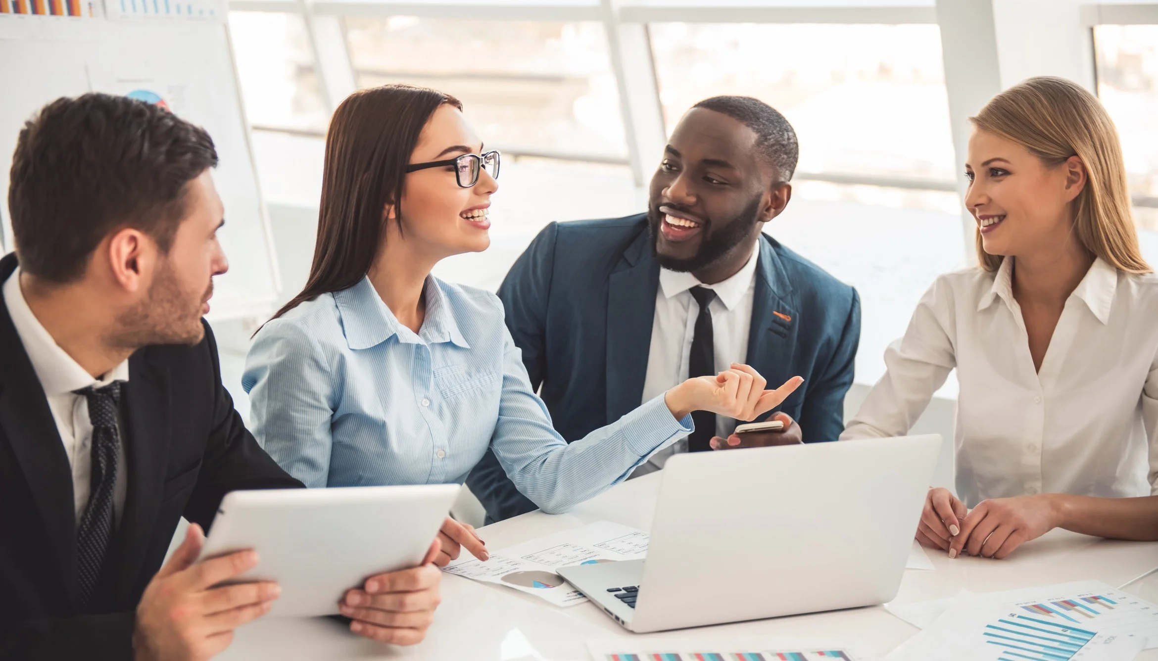 Four diverse business professionals engaged in a lively discussion at a conference table in a modern office. They are smiling, talking, and looking at each other. The table has laptops, charts, and documents.