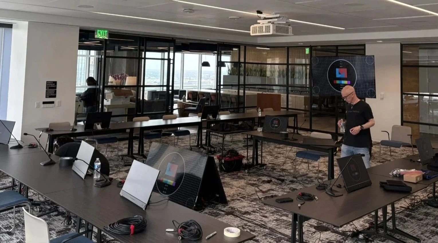 A conference room set up with multiple tables, computer monitors, cables, water bottles, and a person setting up equipment. Large windows and a city view are visible in the background.