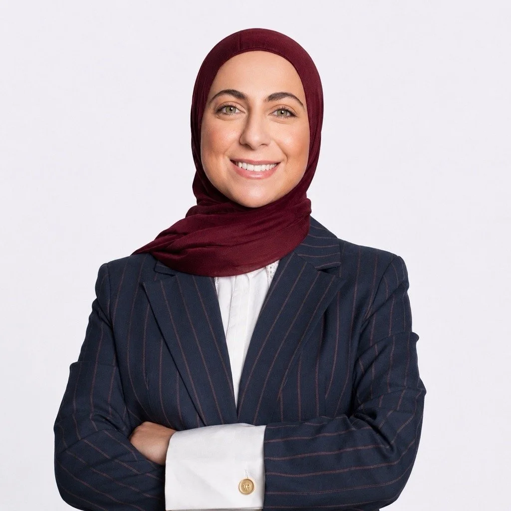 A woman wearing a burgundy hijab and a dark, pinstripe blazer, smiling with arms crossed against a plain white background.