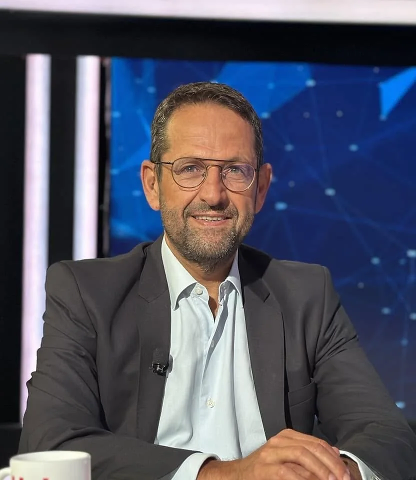 A middle-aged man in a suit and glasses sitting at a table with a coffee mug, smiling at the camera, with a blue digital-themed background.