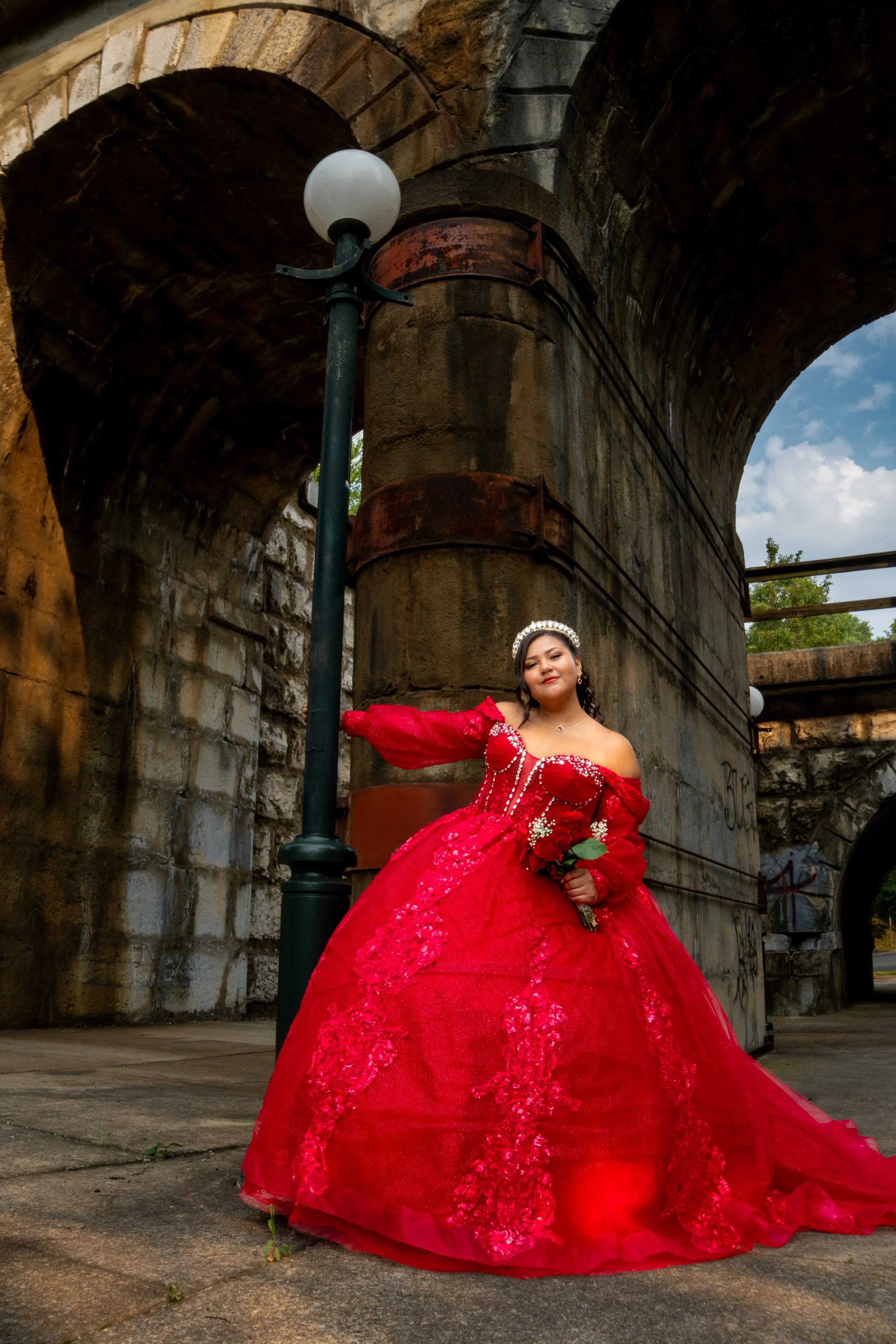 A young woman in a red ball gown with lace and embroidery, wearing a tiara and holding a bouquet of red roses, standing under a stone bridge with a lamp post nearby and a cloudy sky in the background.