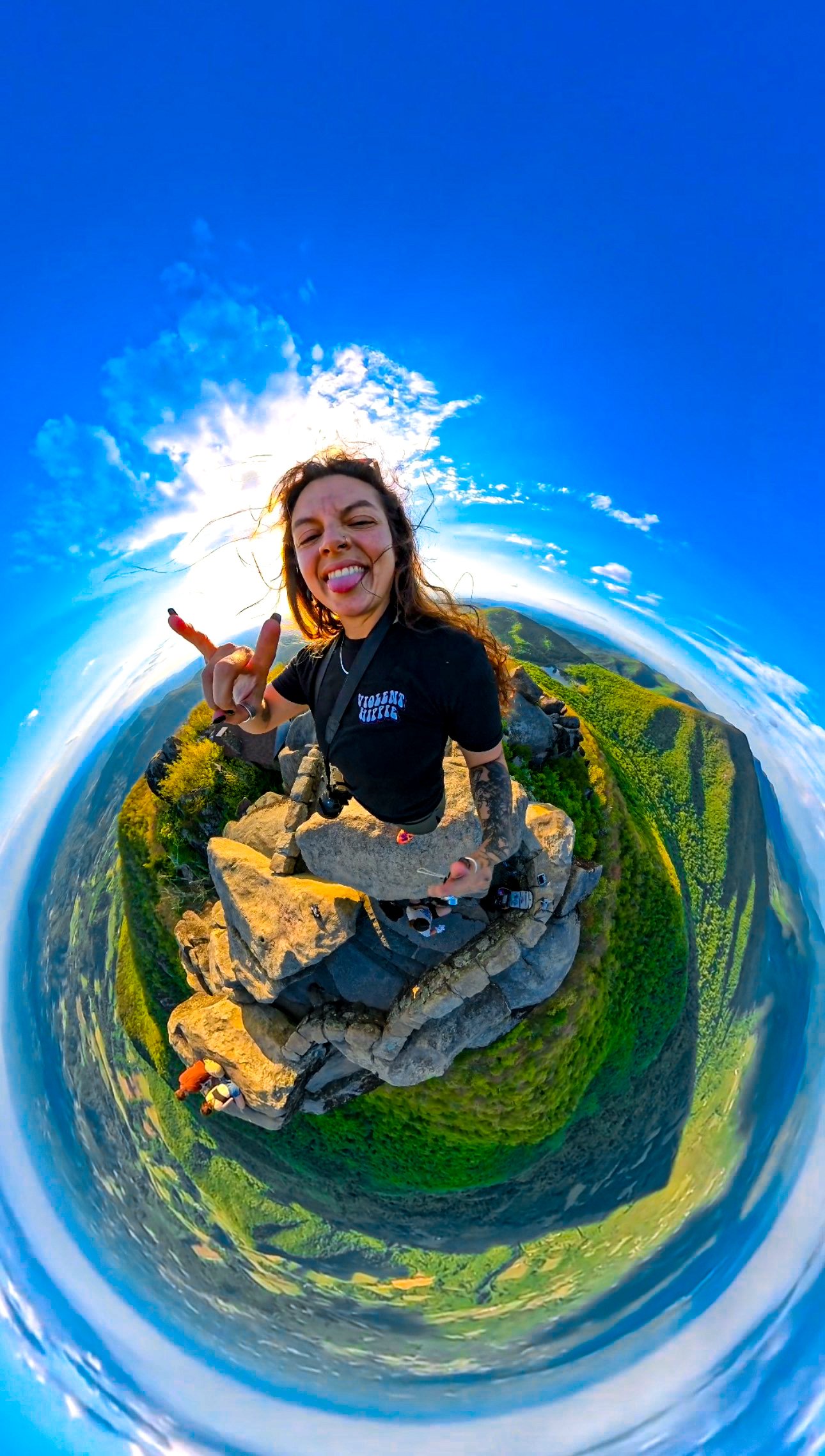 A woman with long hair sticking out her tongue and making a hand gesture, posing on a rocky mountain summit with a panoramic landscape of valleys and hills beneath a blue sky with scattered clouds.