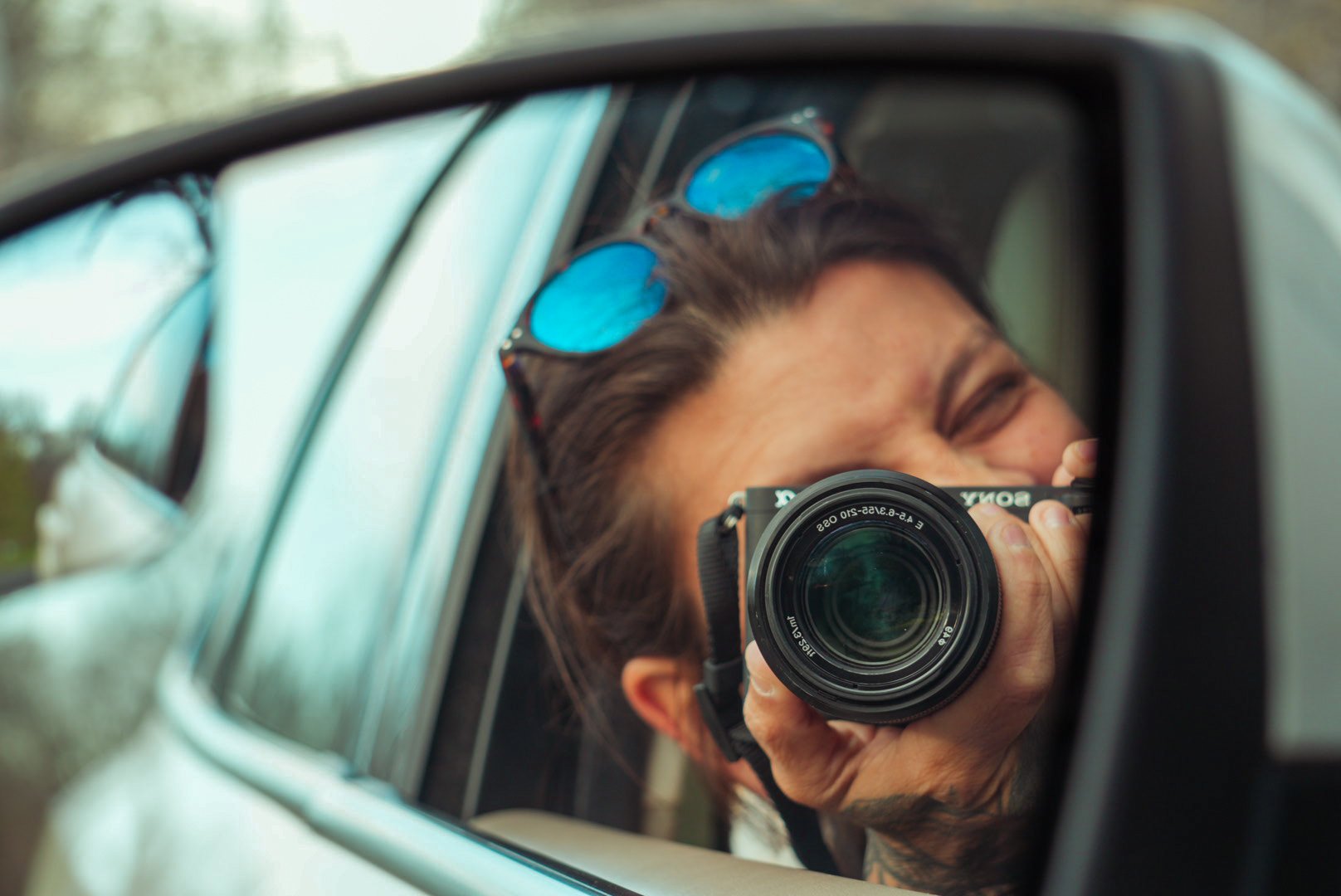 Person with sunglasses on head taking a selfie with a mirrorless camera in the side-view mirror of a car during daytime.