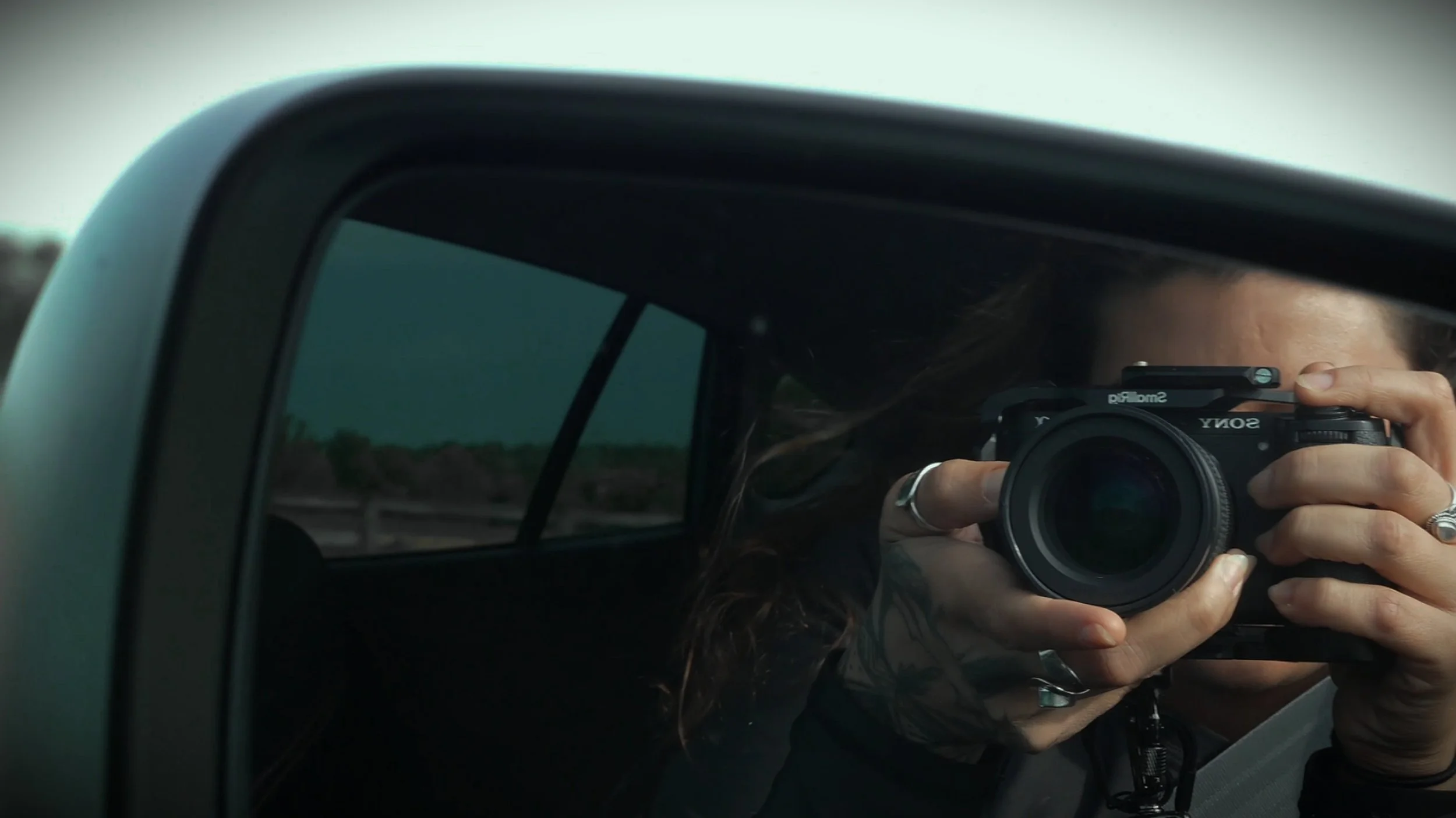 Person taking a mirror selfie with a camera inside a vehicle, with a window showing an outdoor landscape and partly visible face, camera, and hand.