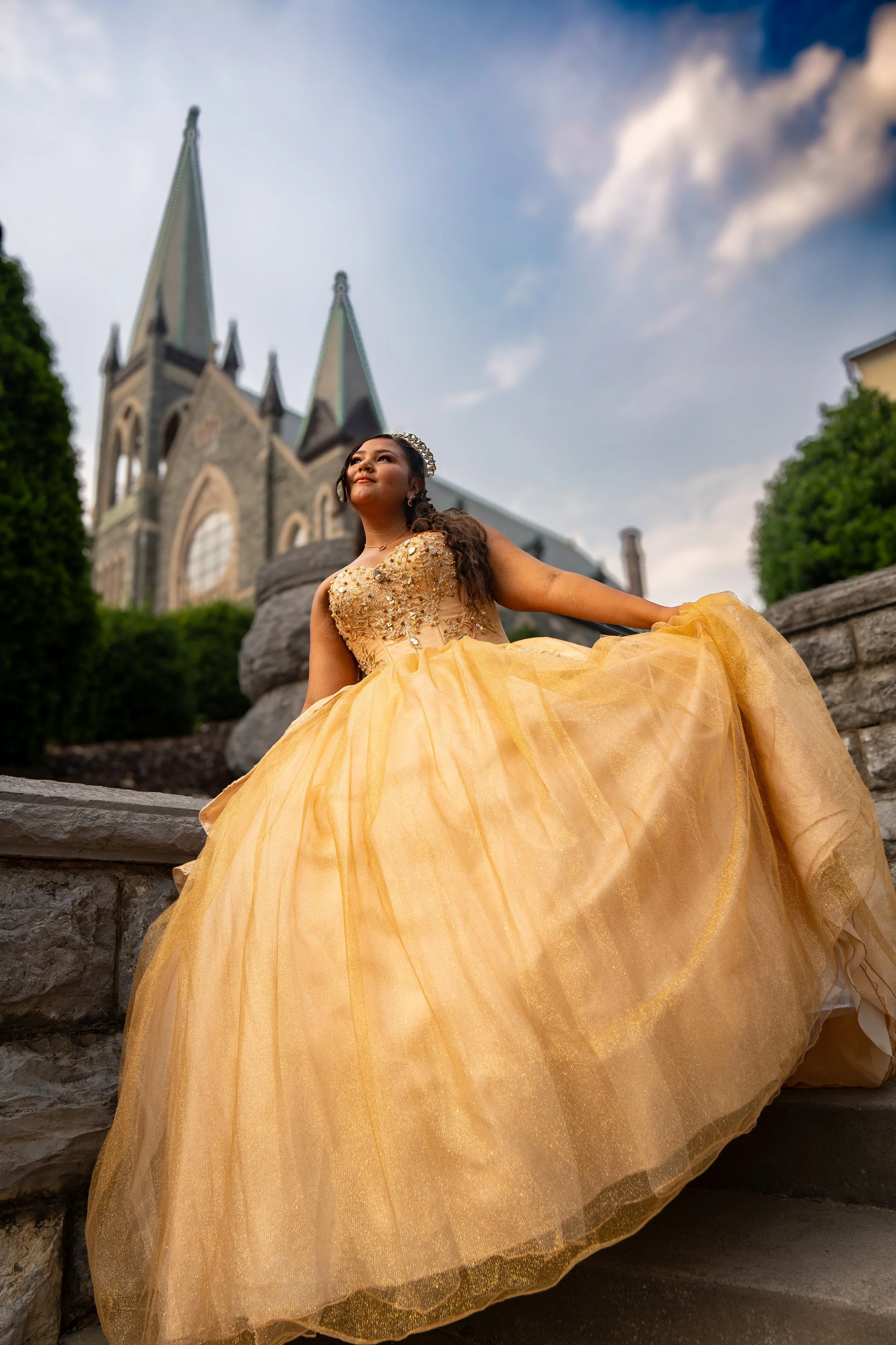 Young woman in a yellow ball gown with embellishments, sitting outdoors on stone steps in front of a historic church with tall spires and a cloudy sky.