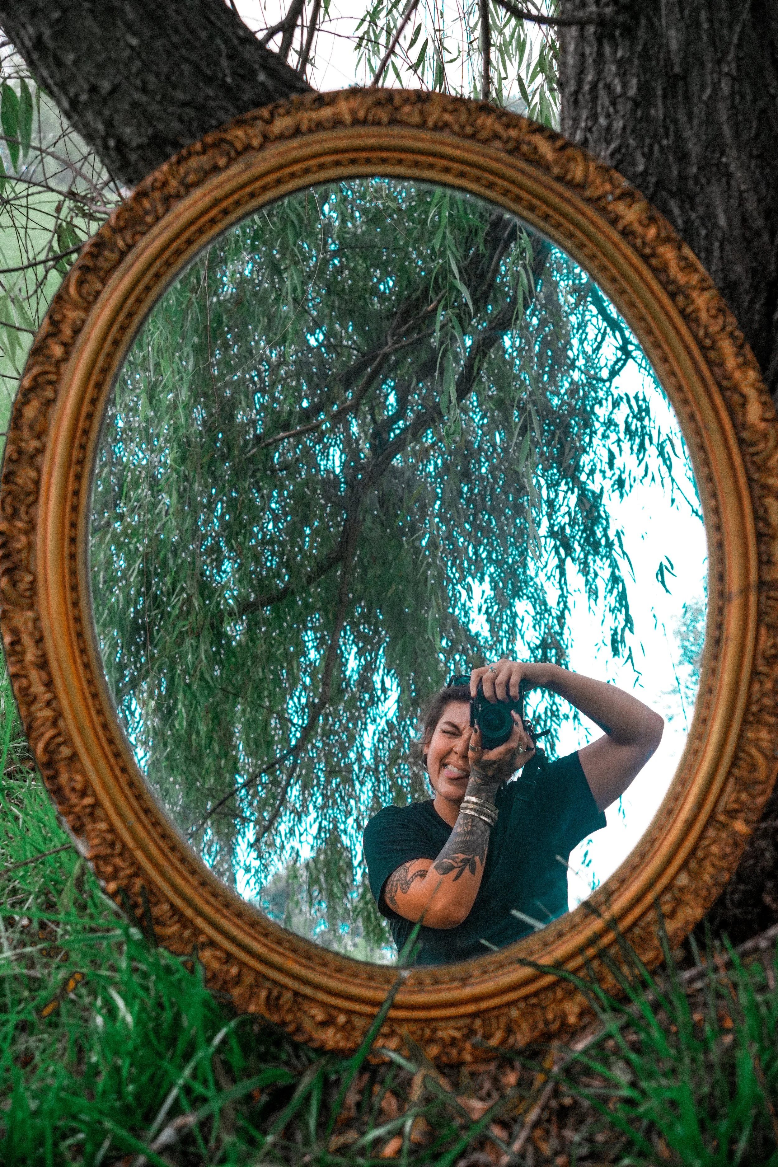 A woman with tattoos on her arm is taking a photo of herself in a decorative round mirror outdoors among trees and green grass, with her tongue slightly out, and holding a camera in front of her face.