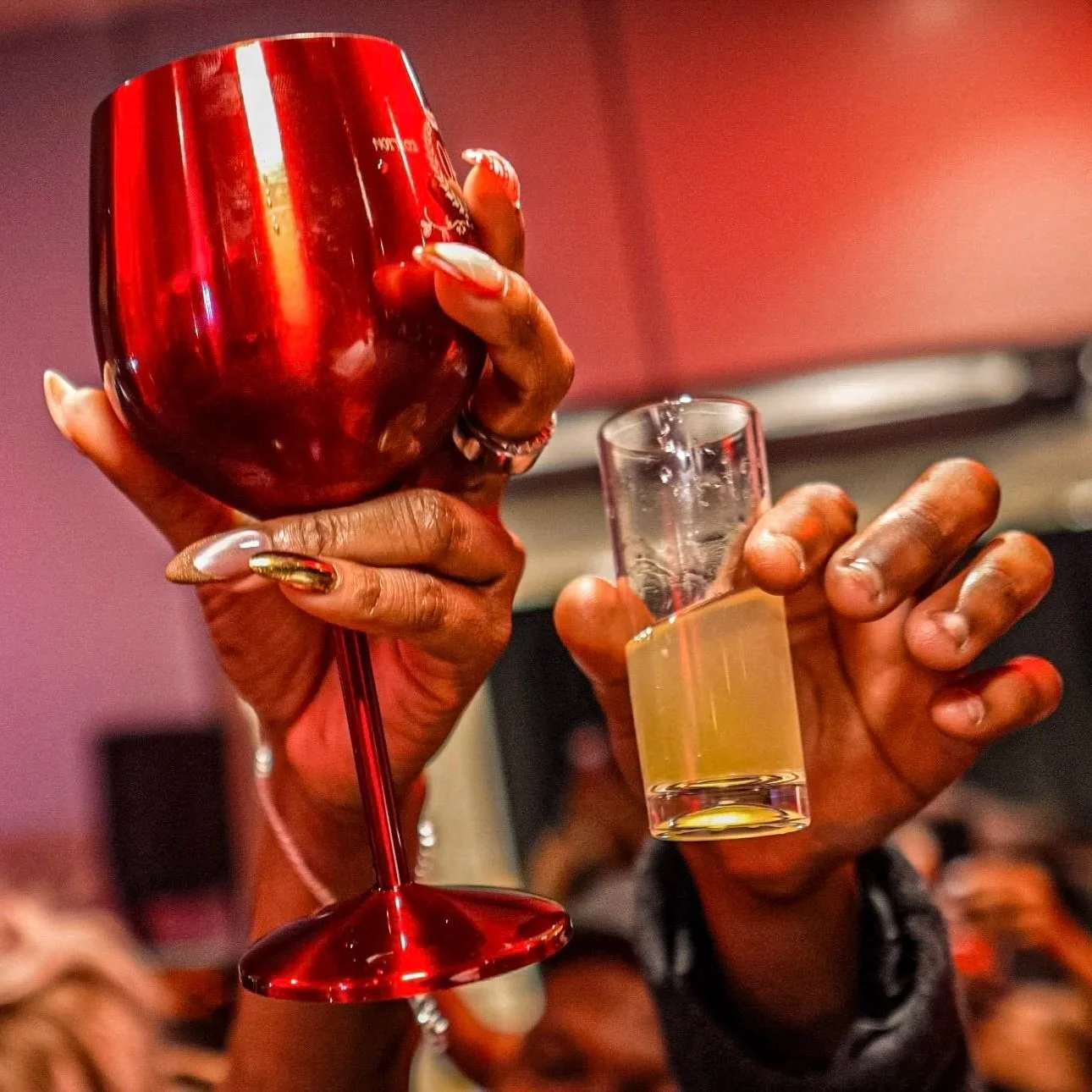 Person holding a red wine glass in one hand and a shot glass filled with a clear beverage in the other, celebrating at a social gathering.