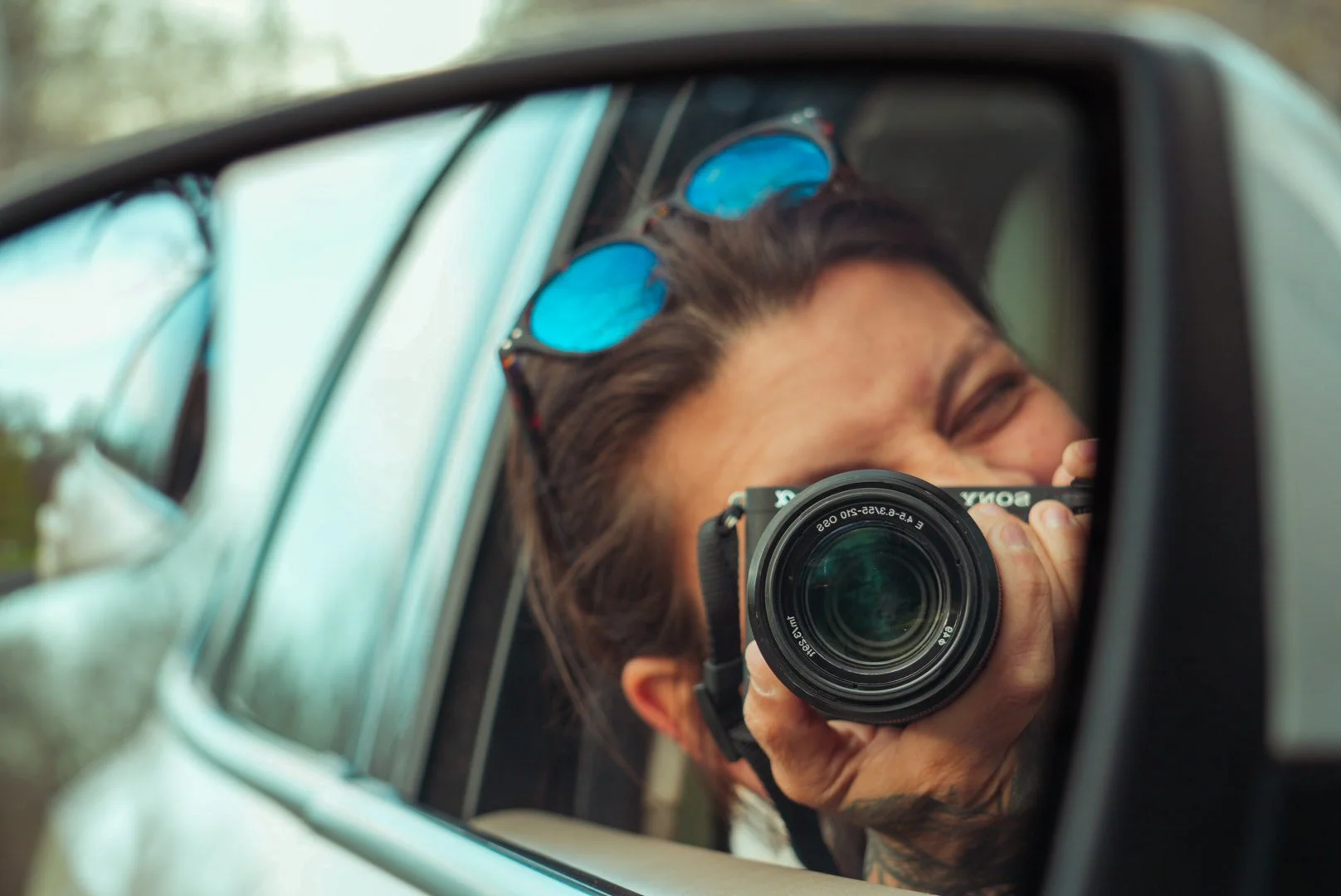 Person taking a photo with a Sony camera reflected in a car's side mirror, wearing sunglasses on their head, outdoors on a cloudy day.
