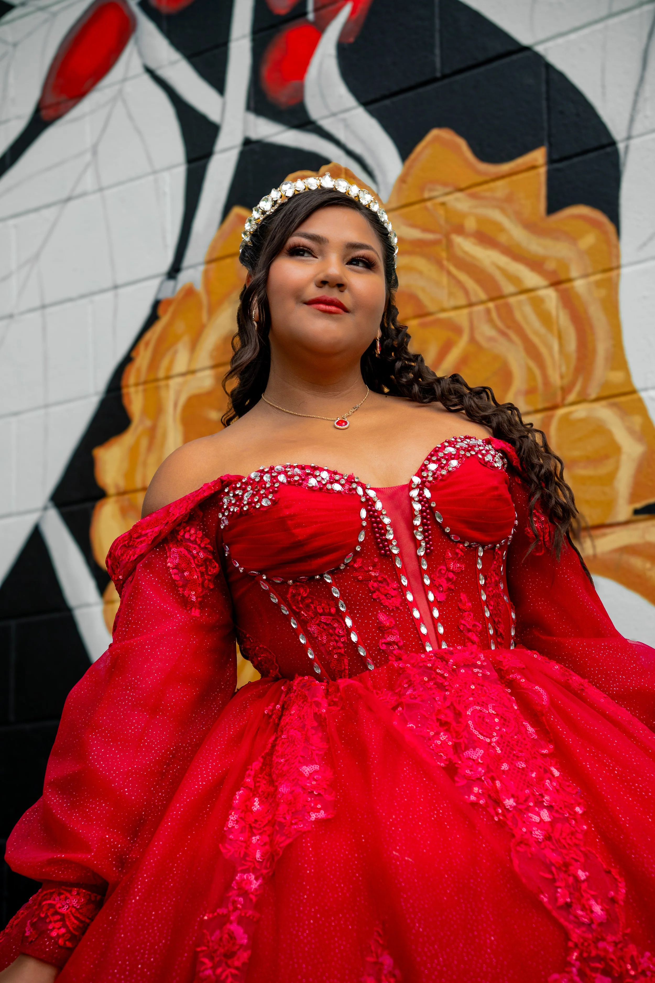 A woman in a vibrant red dress with rhinestone embellishments, wearing a pearl and gemstone headband and a matching jewelry set, standing in front of a colorful mural painted on a brick wall.