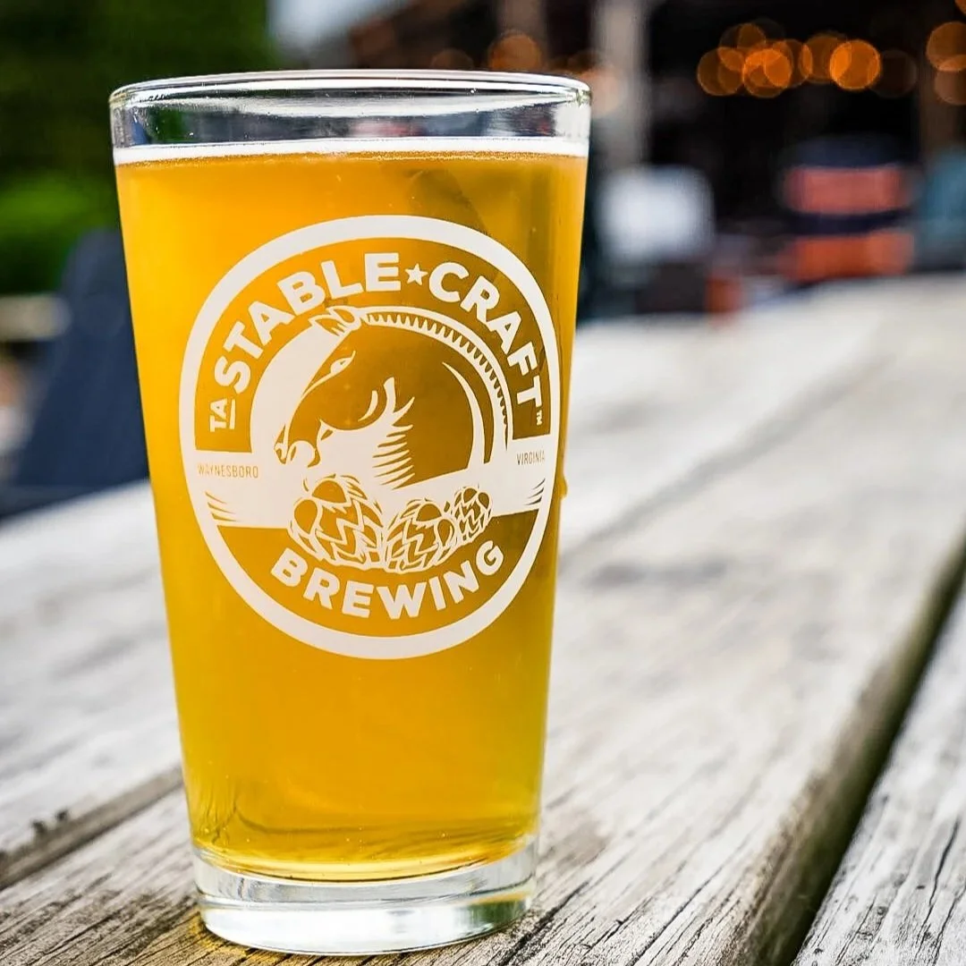 A pint glass of beer on a wooden surface with the logo of Stable Craft Brewing on it, outdoors with blurry lights and background.