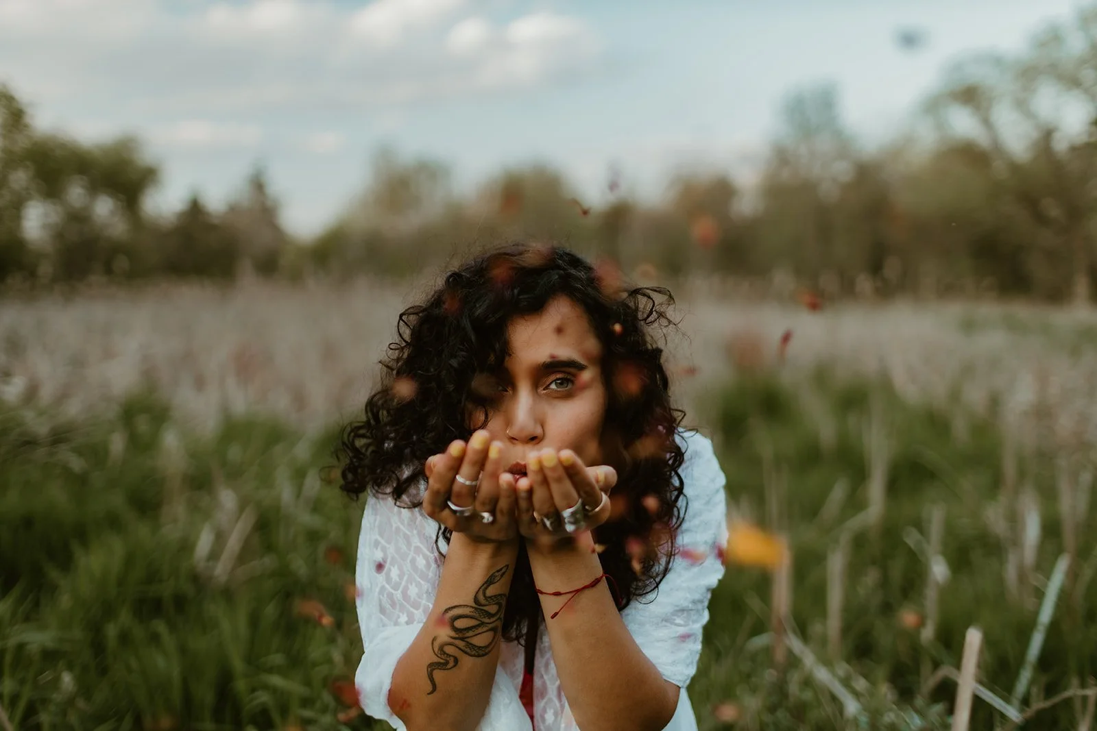 A woman with curly hair blowing flower petals outdoors in a grassy field.