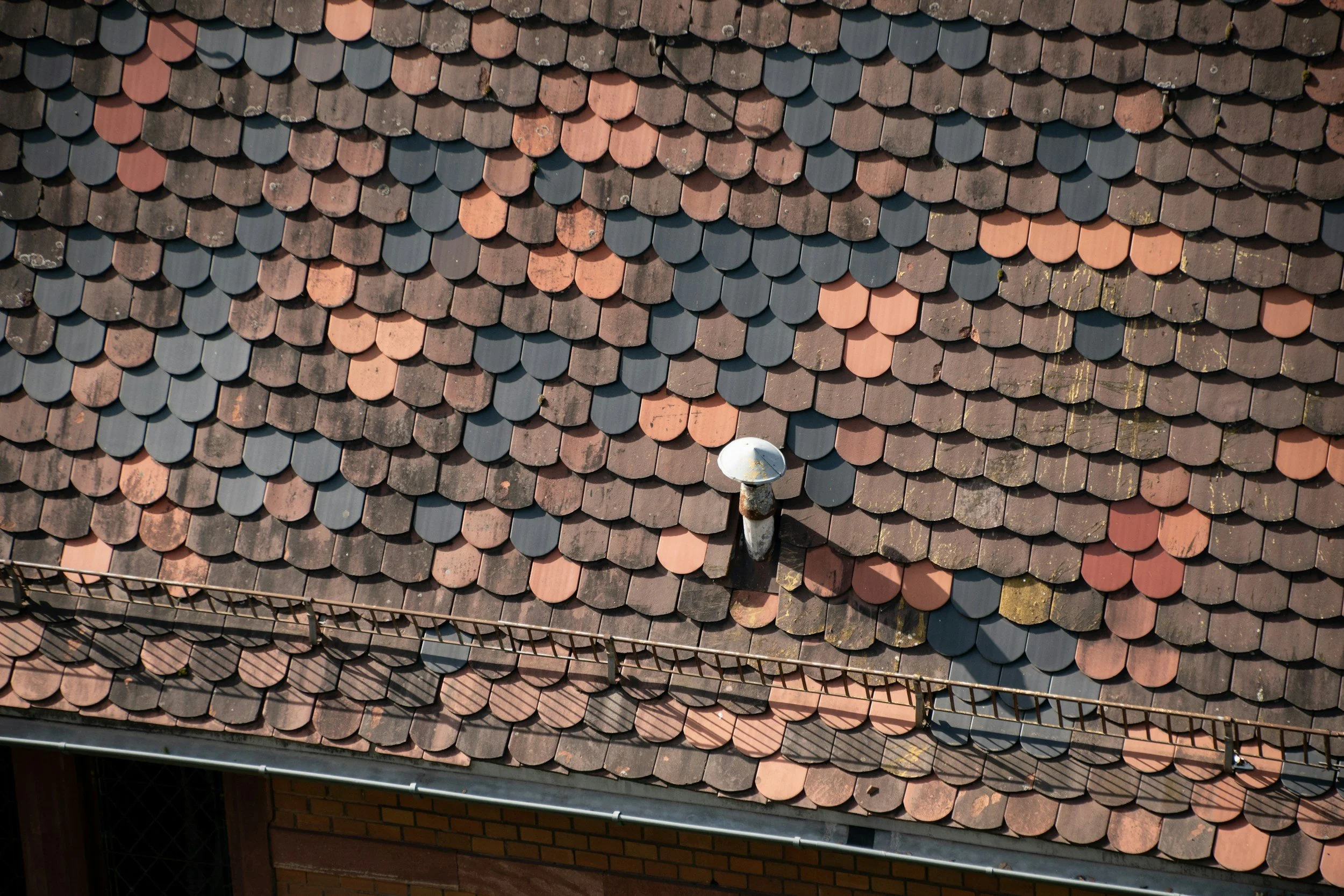 A rooftop with overlapping brown and gray tiles, with a vent pipe and security fencing visible at the lower part of the image.