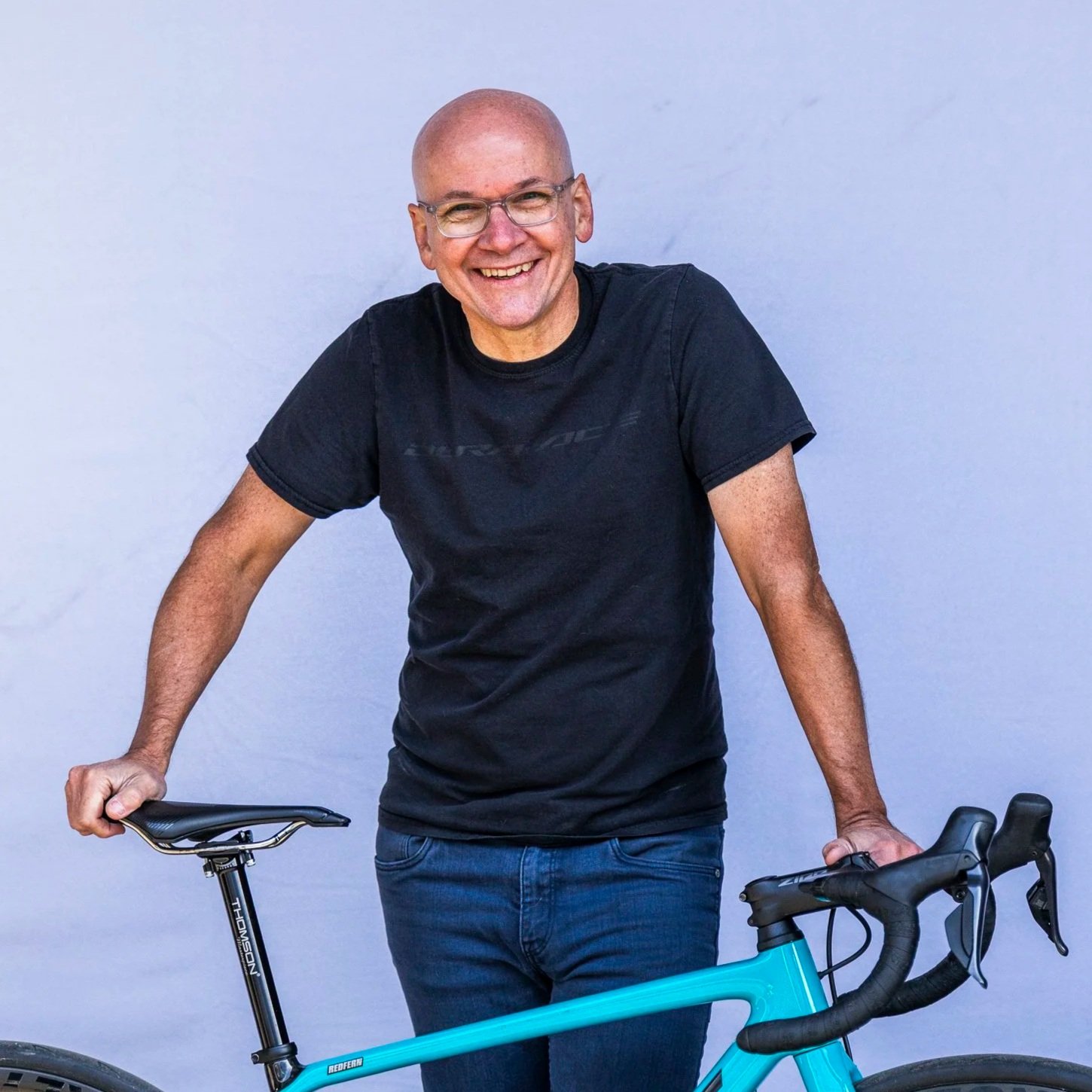 A smiling middle-aged man with glasses wearing a black t-shirt and jeans, standing with one hand on a bicycle against a plain light-colored background.