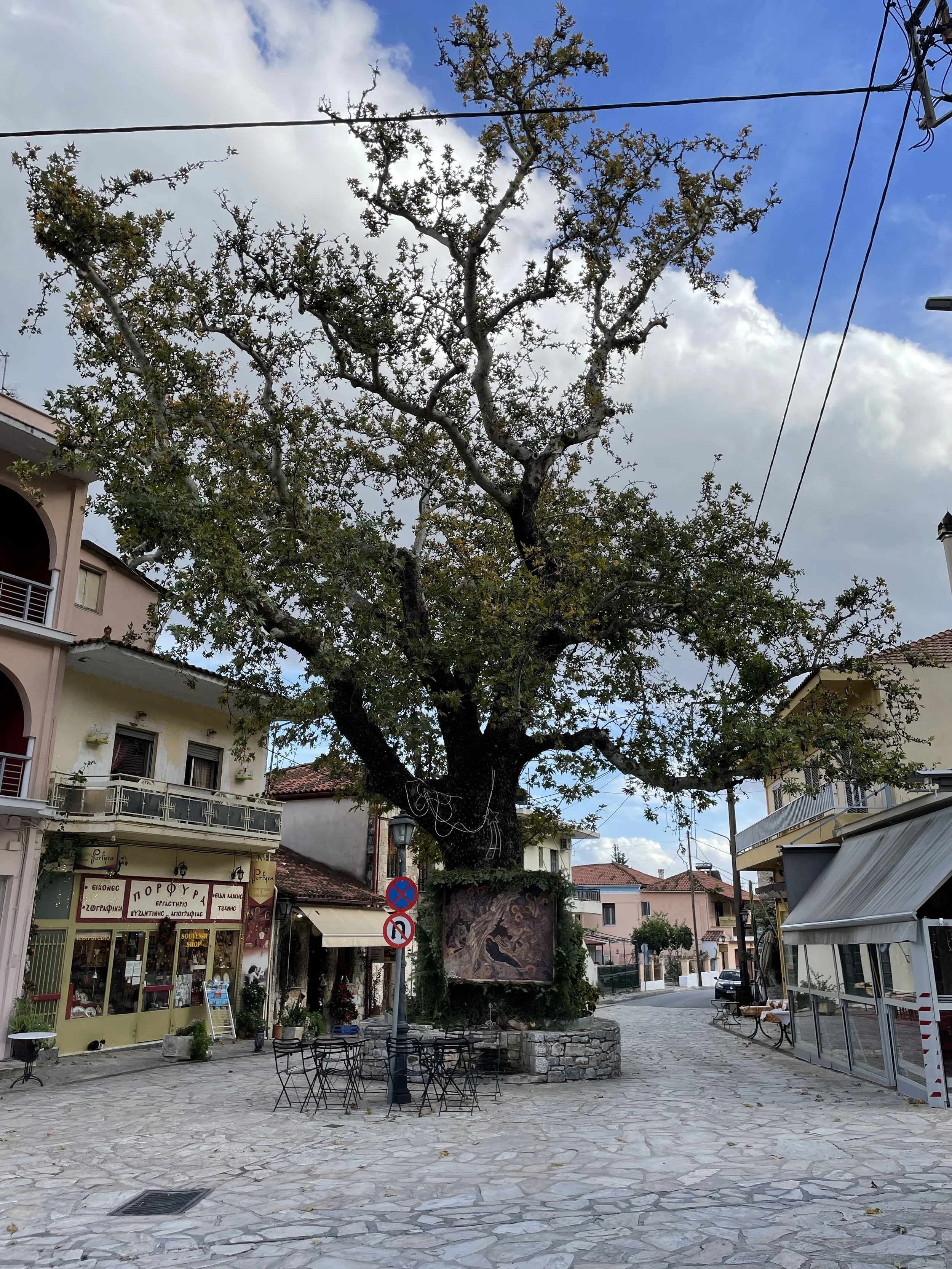 A small town square with a large tree in the center, surrounded by buildings, shops, and cafes. There are chairs and tables arranged around the tree, and a street with parked cars and signs. The sky is partly cloudy with patches of blue visible.