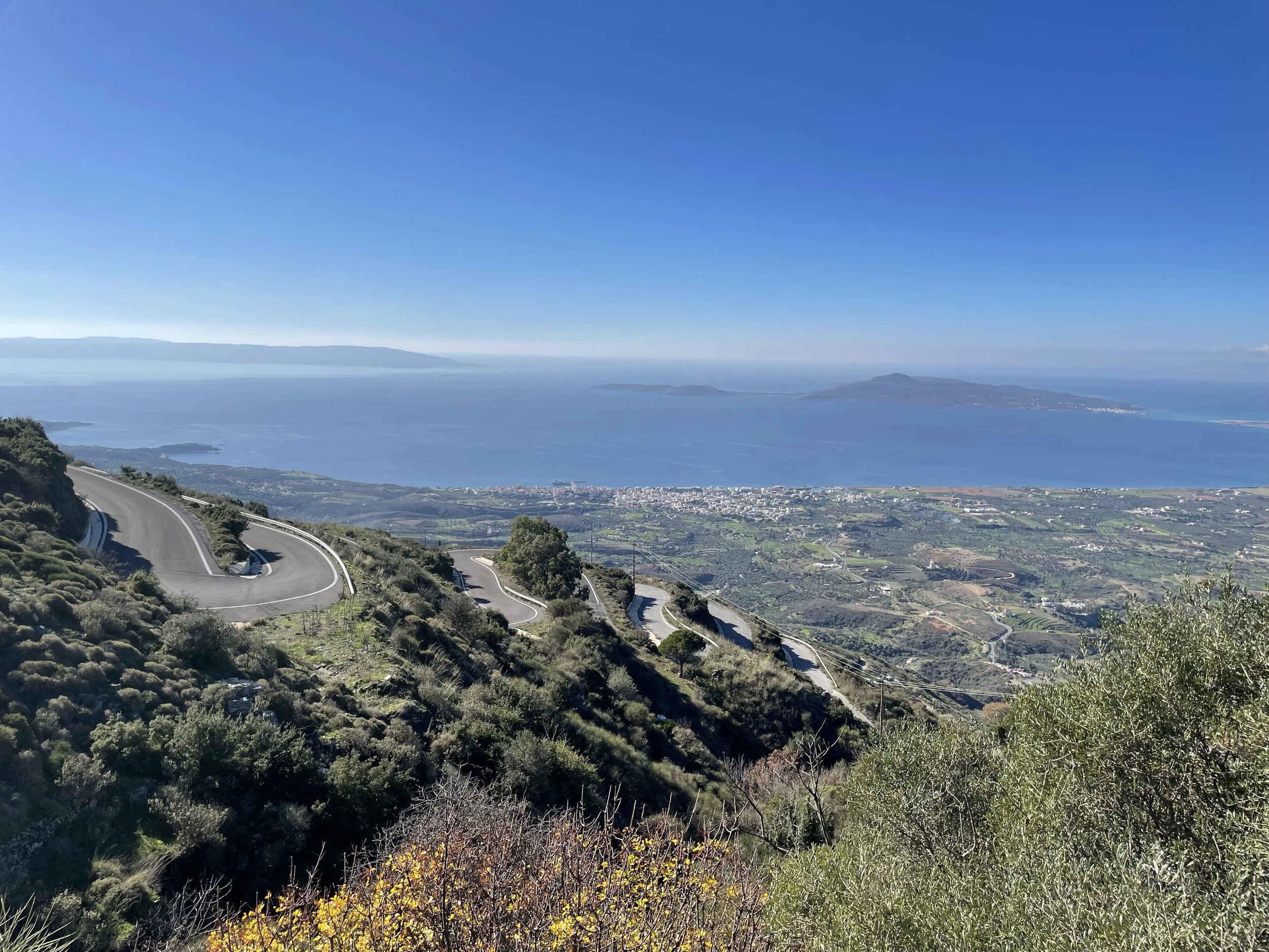 Winding road on a hillside overlooking a large body of water and distant islands under a clear blue sky.