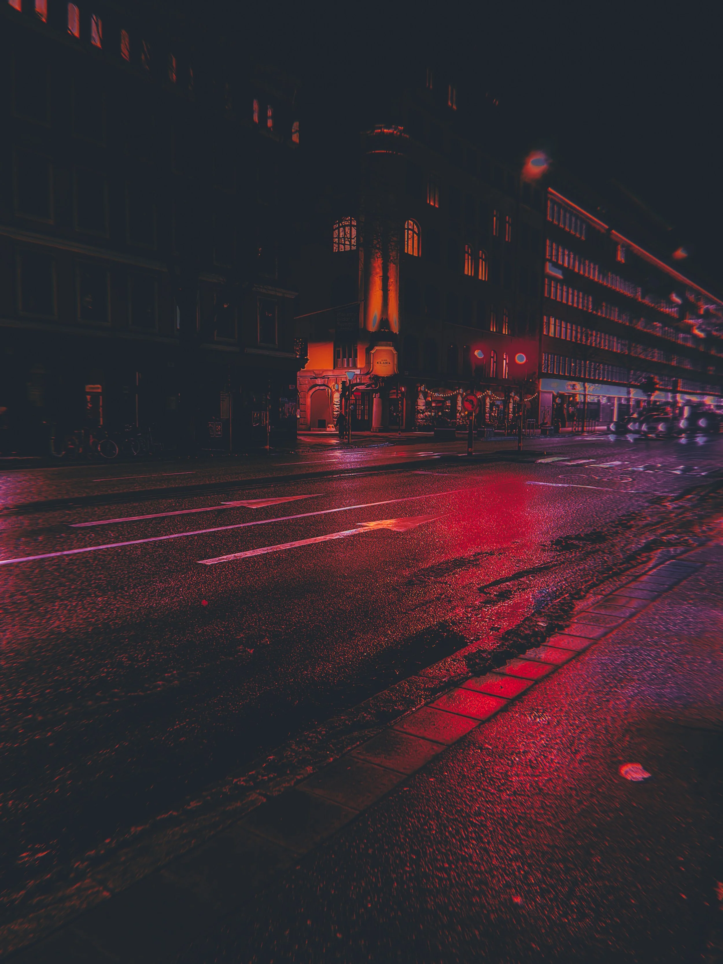 Nighttime city street scene with wet pavement reflecting red and purple lights from nearby buildings, illuminated windows, and streetlights.