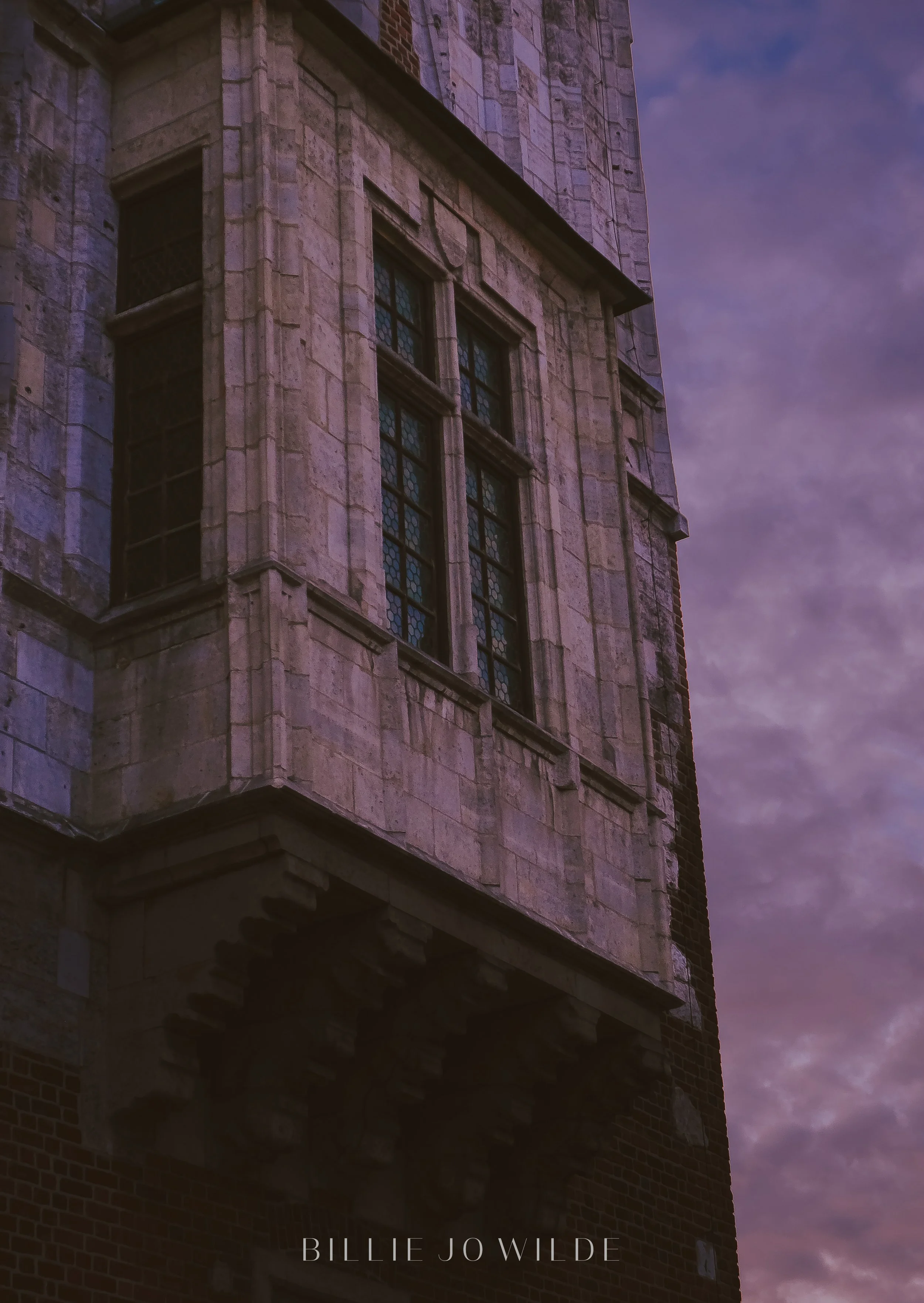 Close-up of a historic stone building with window frames, part of a taller structure, under a cloudy purple evening sky. Text overlay says 'Billie J Wilde' at the bottom.