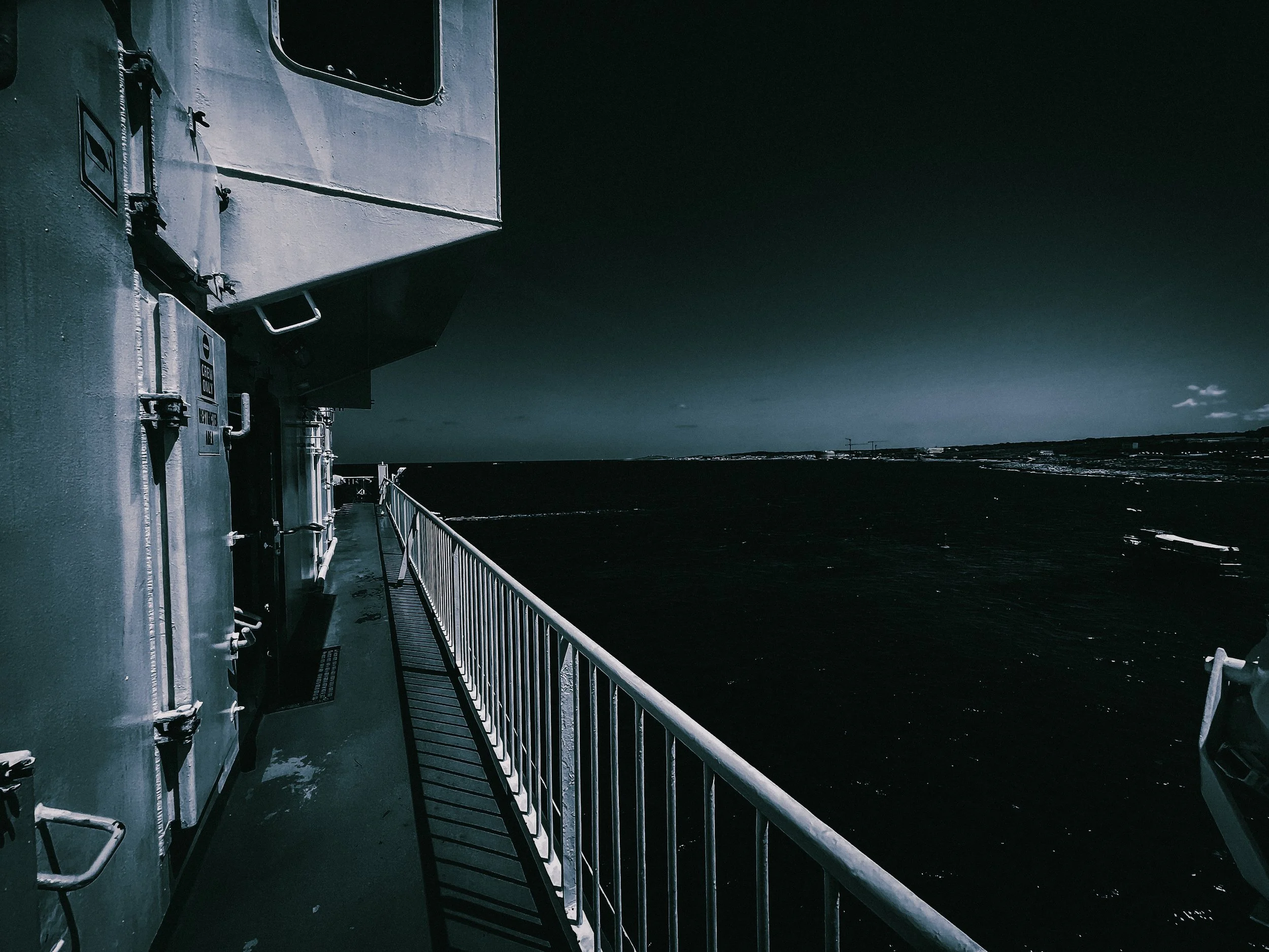View from the deck of a ship, showing industrial equipment and a metal railing, with a dark ocean and a distant horizon under dramatic clouds in the background.