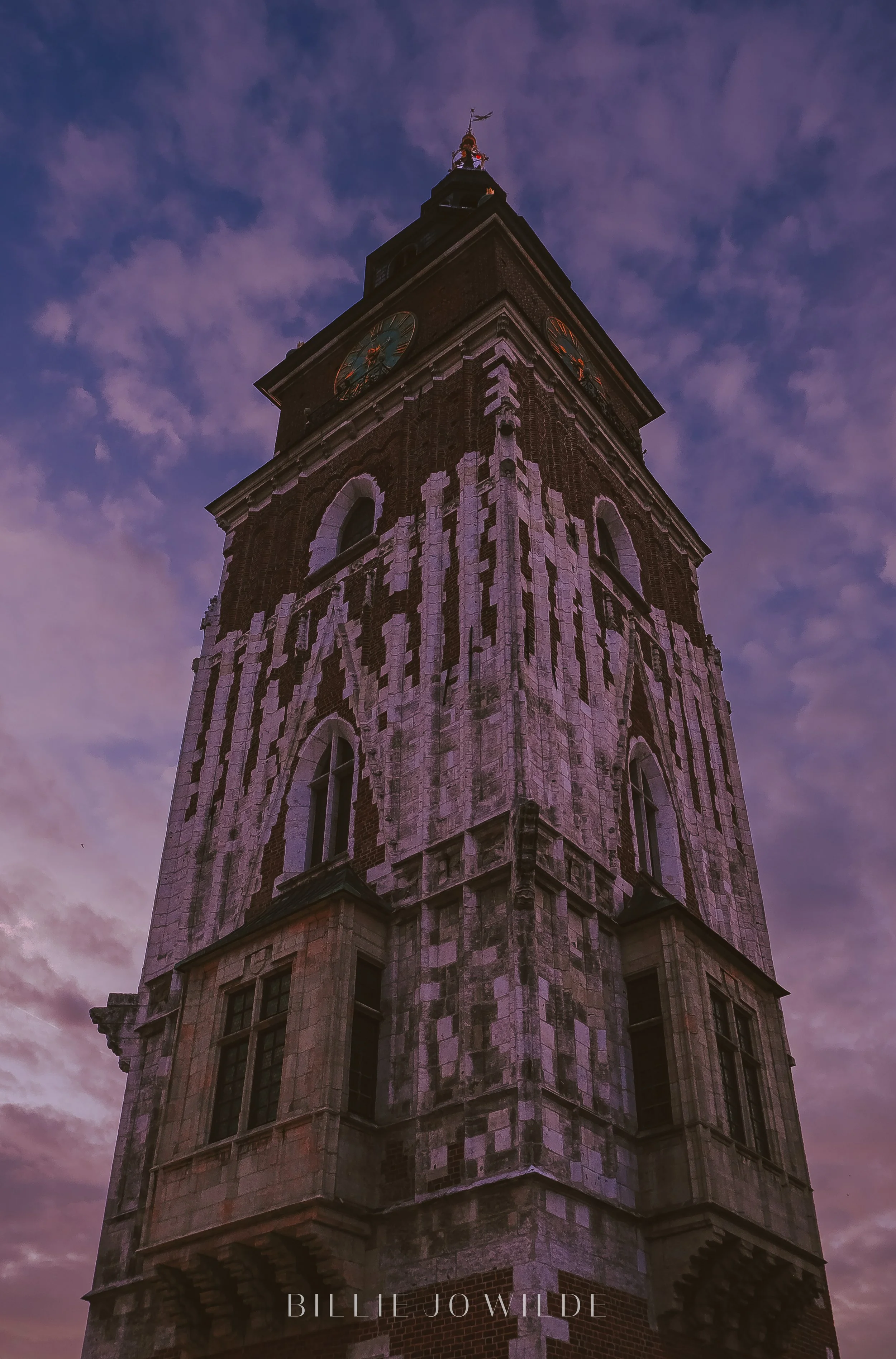 A tall brick clock tower against a purple cloudy sky at dusk, with a weather vane at the top.