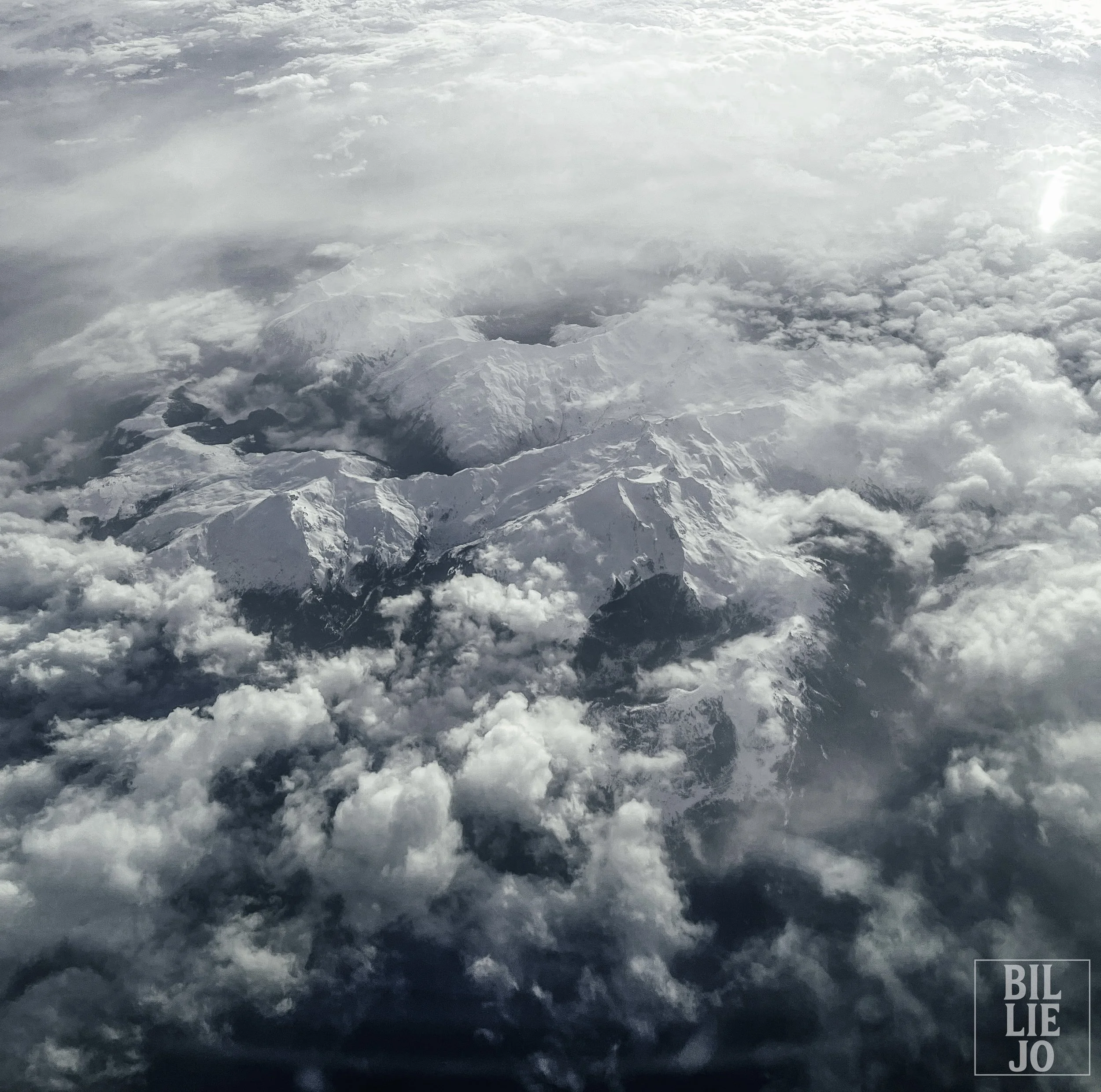 Snow-covered mountain peaks surrounded by clouds with bright sunlight in the sky.
