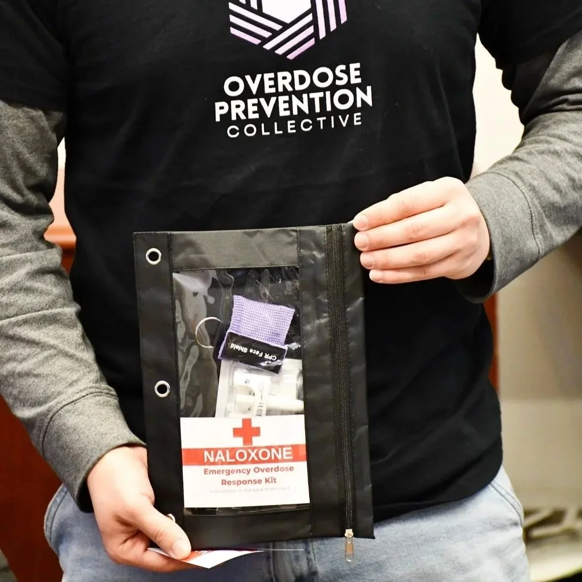 Person holding a black emergency overdose response kit with naloxone, inside a clear plastic pocket, wearing a black shirt with white text about overdose prevention