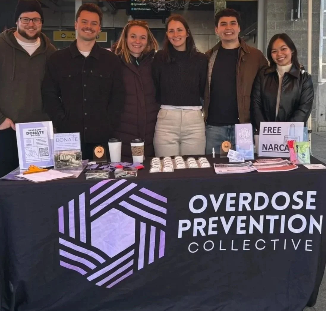 Six people standing behind a table at an overdose prevention event, with signs, informational pamphlets, and supplies on the table.