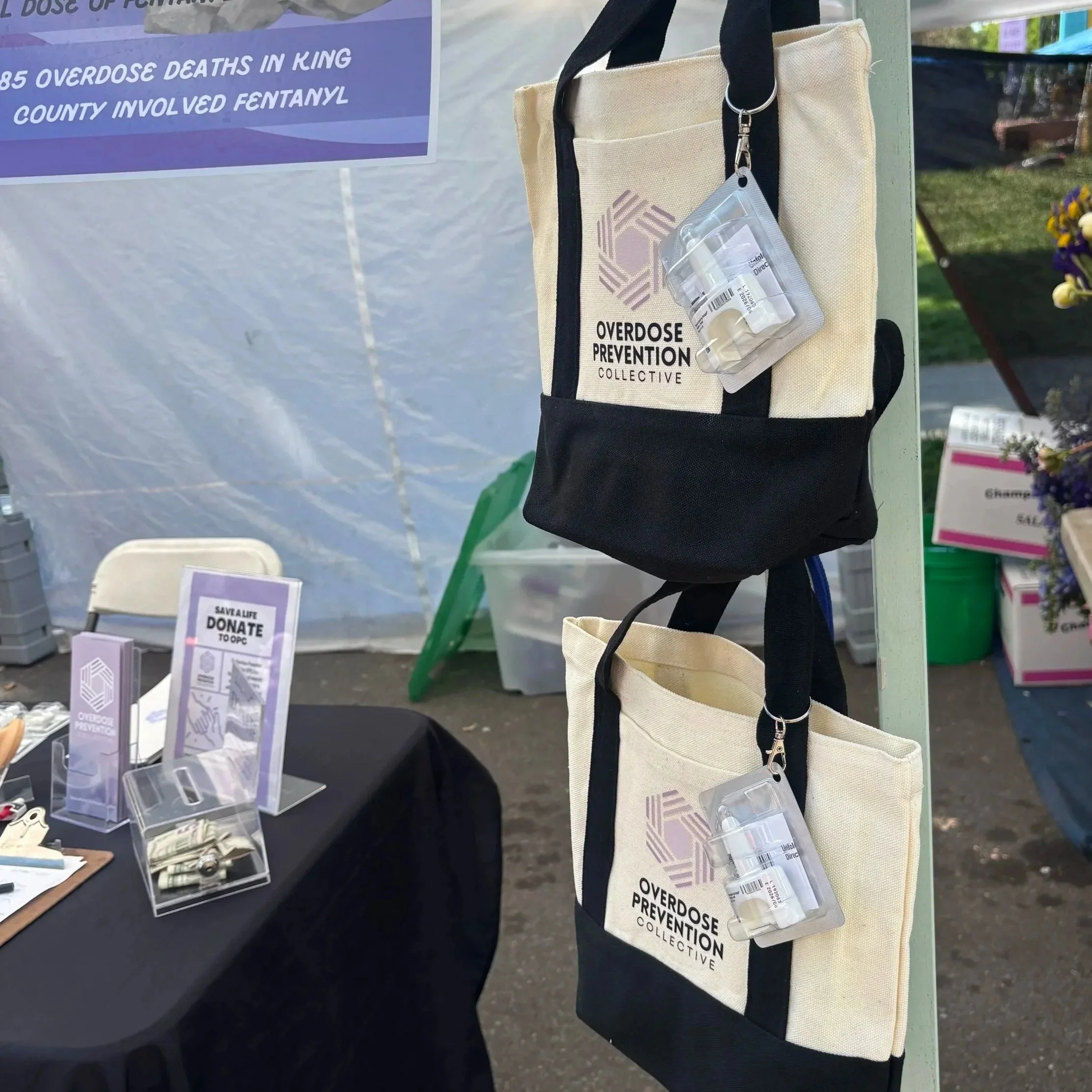 A beige shoulder bag with a logo and the words "Overdose Prevention Collective" printed on it, hanging from a strap with a clear plastic container of pills attached. Part of a sign in the background mentions fentanyl and overdose deaths.