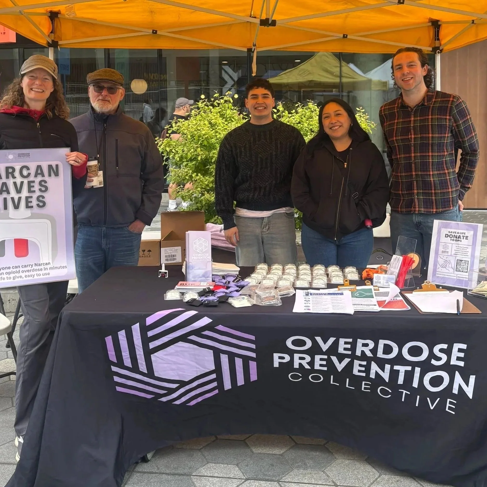 Group of five people standing behind a table at a public event, under a yellow tent, with signs and informational materials promoting overdose prevention and harm reduction.