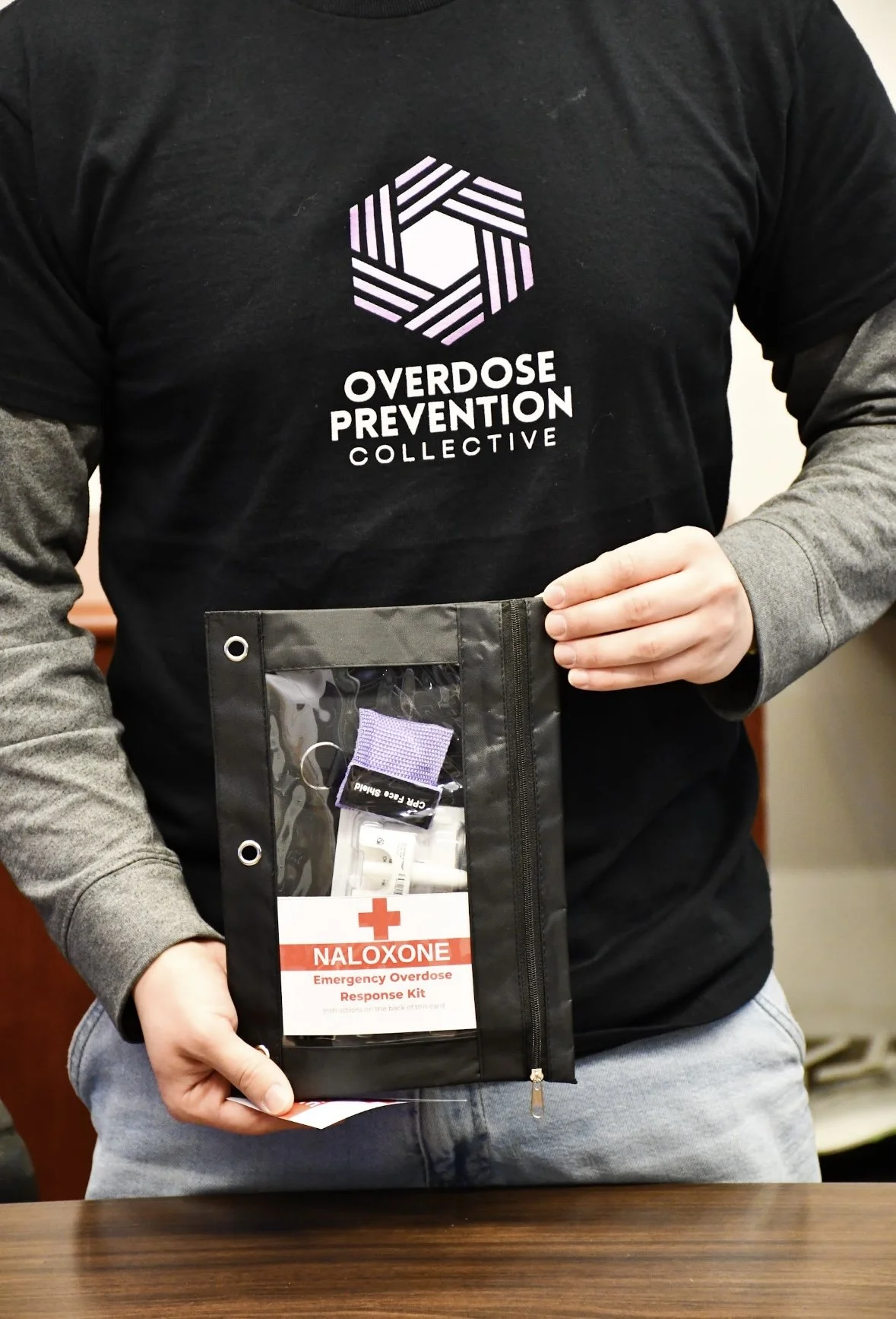 Person wearing a black t-shirt with the logo and text 'Overdose Prevention Collective' and holding an emergency overdose response kit labeled for Naloxone.