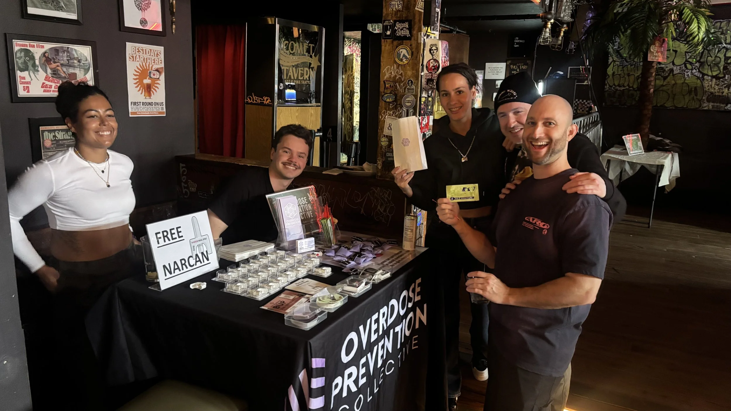 Group of five people smiling and posing for the camera at a prevention event booth, with pamphlets, signs, and a table displaying Narcan supplies, inside a dimly lit room with posters and graffiti on the walls.
