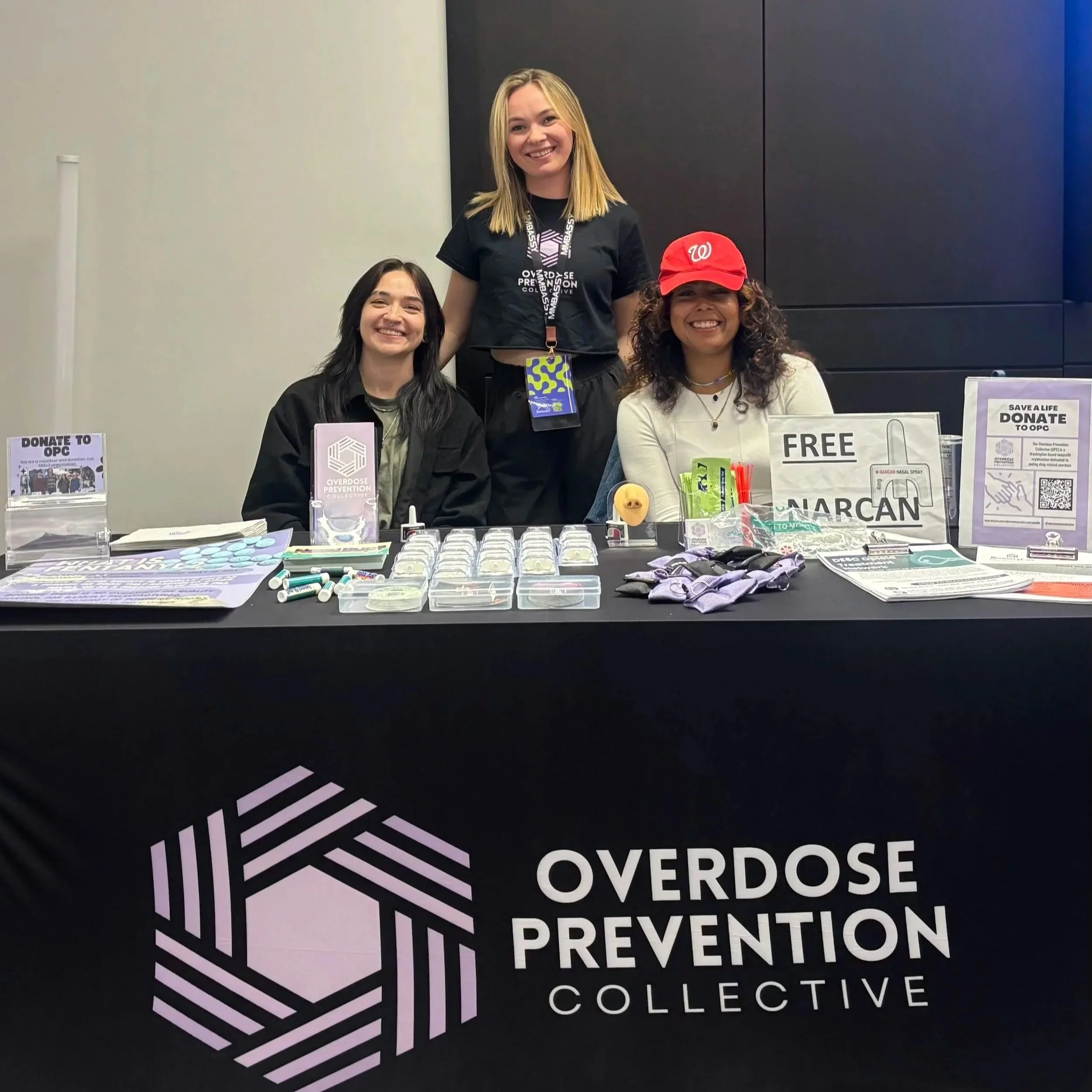 Three women smiling at a table with printed materials and informational displays about overdose prevention, overdose prevention collection, and drug overdose prevention.