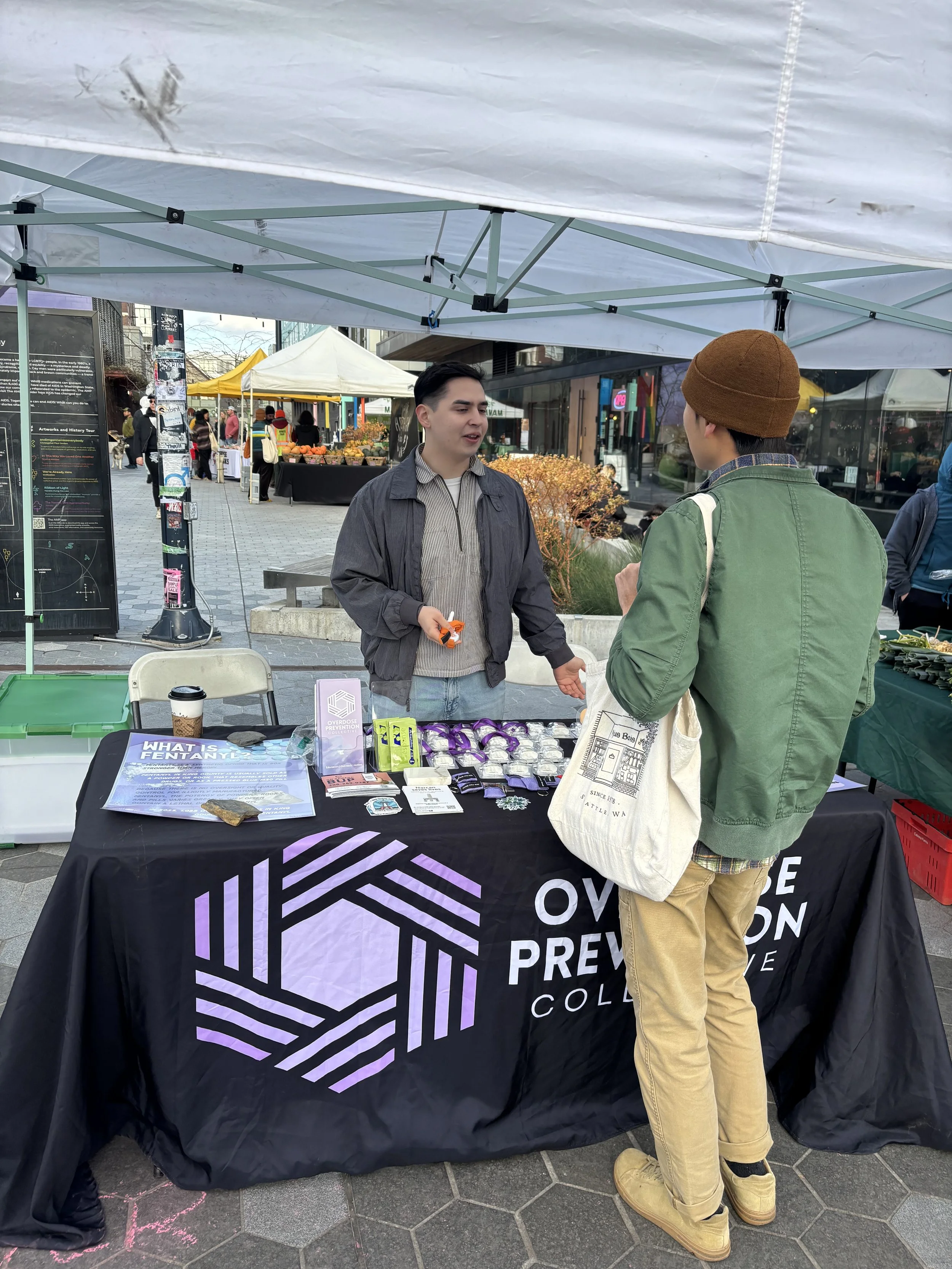 Two people are talking at an outdoor table with a black tablecloth that has purple and white geometric text reading 'Overdose Prevention Coalition.' One person is wearing a gray jacket and striped shirt, holding an orange object. The other person is wearing a green jacket, beige pants, tan boots, and a brown beanie, carrying a white tote bag. There are several informational pamphlets on the table about fentanyl and overdose prevention. In the background, there is a street market with tents and people walking around.
