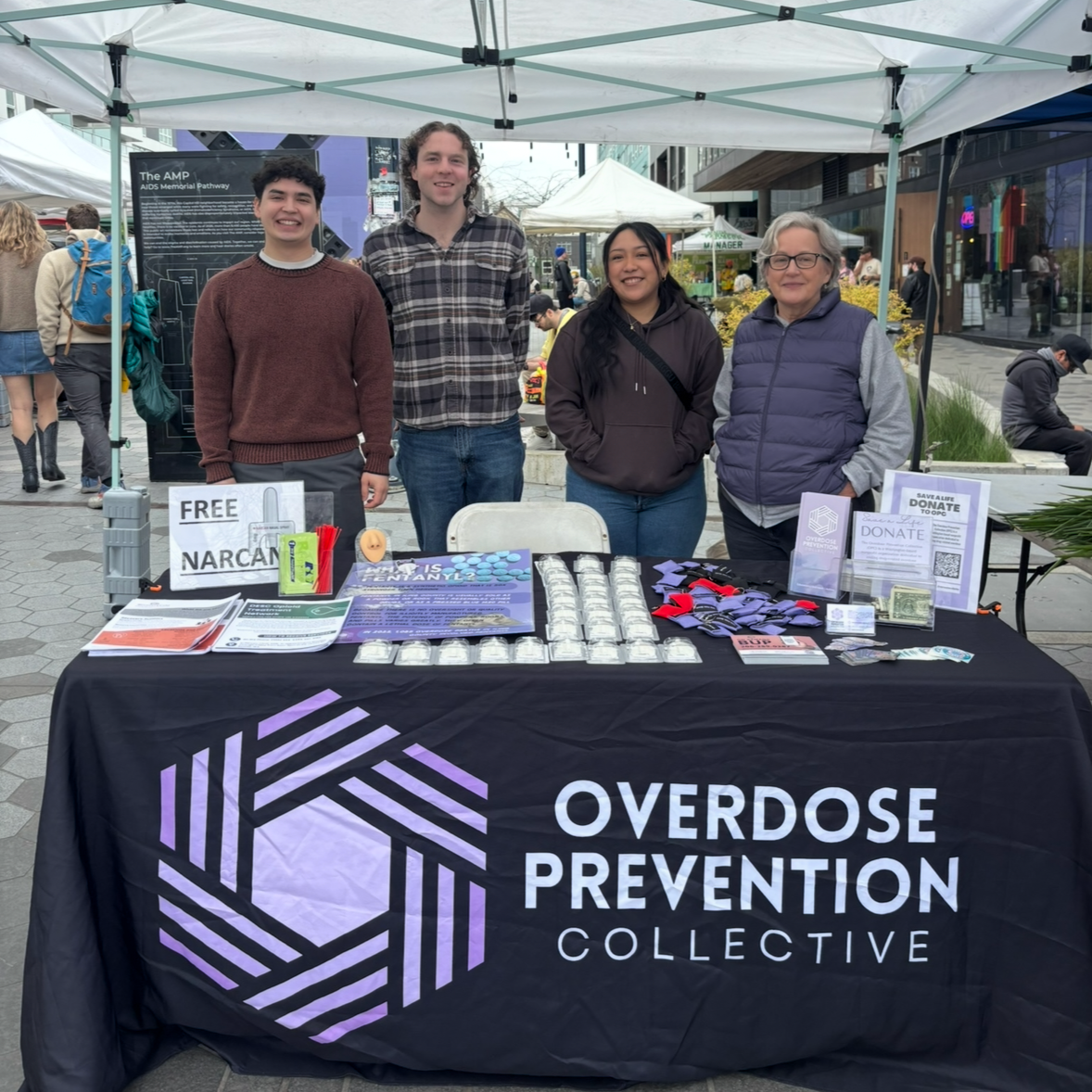 Four people standing behind an overdose prevention table at an outdoor event, with informational materials, free Narcan, and donation signs displayed on the table