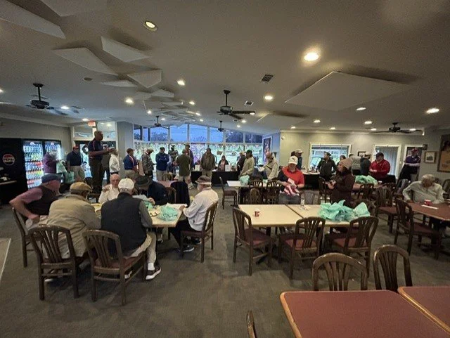 People gathered in a restaurant or event space, some seated at tables and others standing near a bar area with a large window.