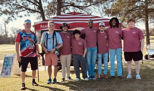 Group of seven people standing outdoors under a red and white striped canopy, smiling for a photo, with trees and a golf cart in the background.