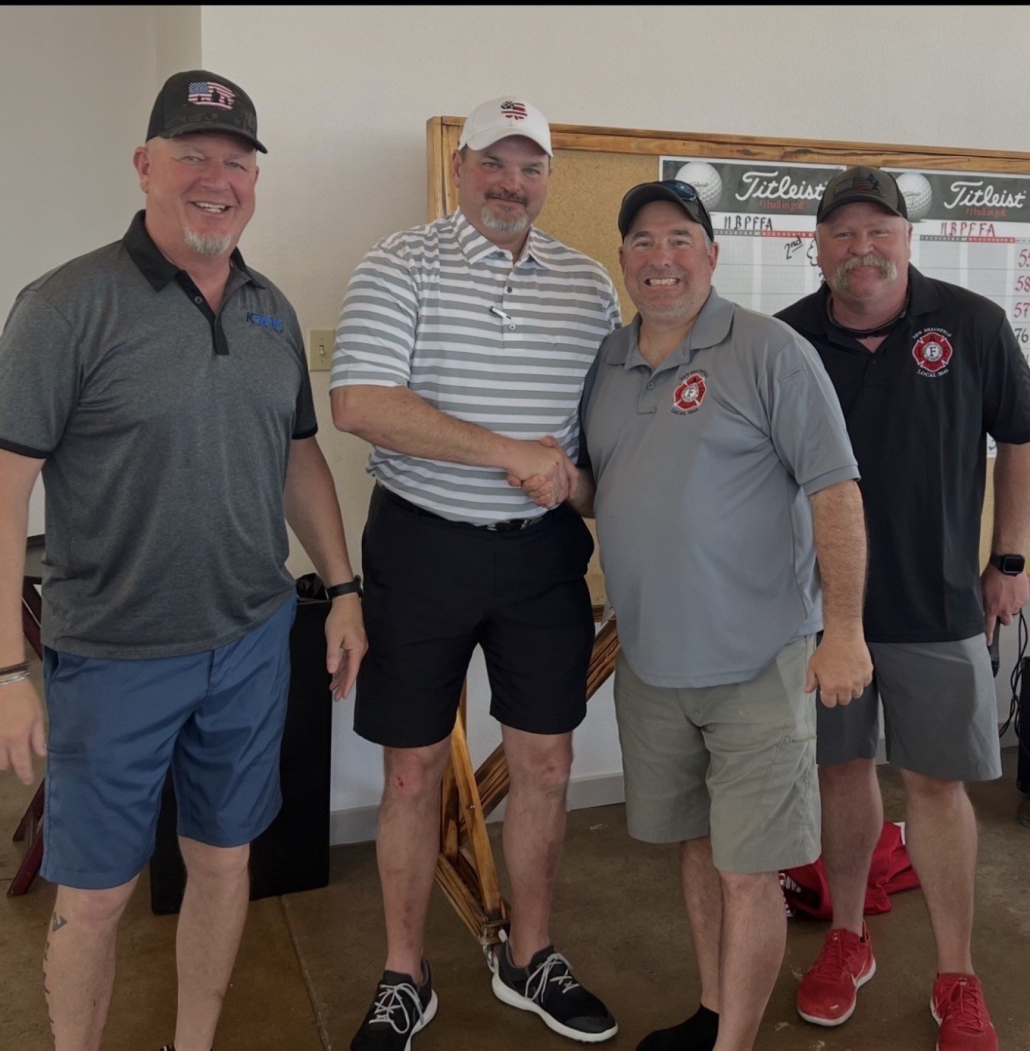 Four men at a golf event, two are shaking hands and smiling, with a golf leaderboard in the background.