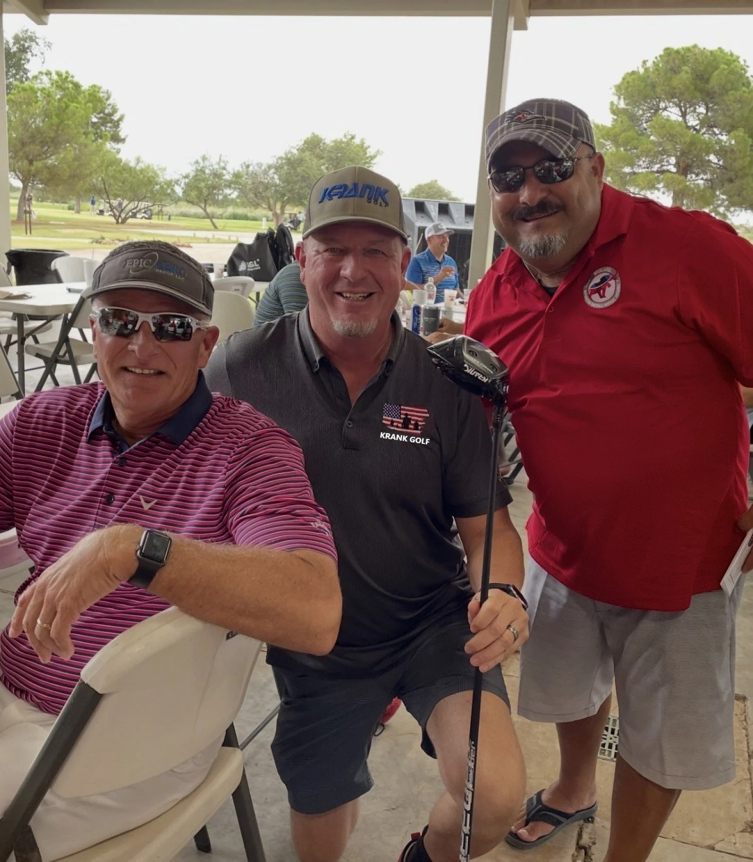 Three men at a golf event, two standing and one sitting, with trees and a golf course in the background. They wear golf hats, sunglasses, and casual golf attire.