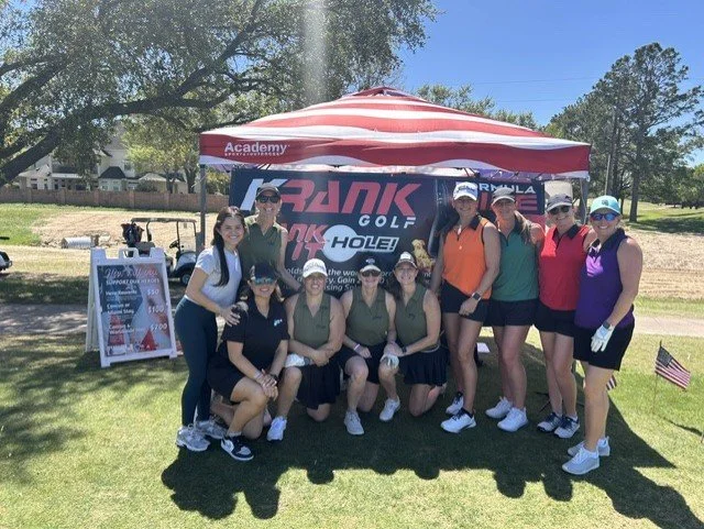 A group of women on a golf course posing for a photo in front of a red and white canopy. They are dressed in athletic golf attire, with some wearing visors or caps. There is a signboard with prices and a small American flag in the background, along with trees and a clear blue sky.