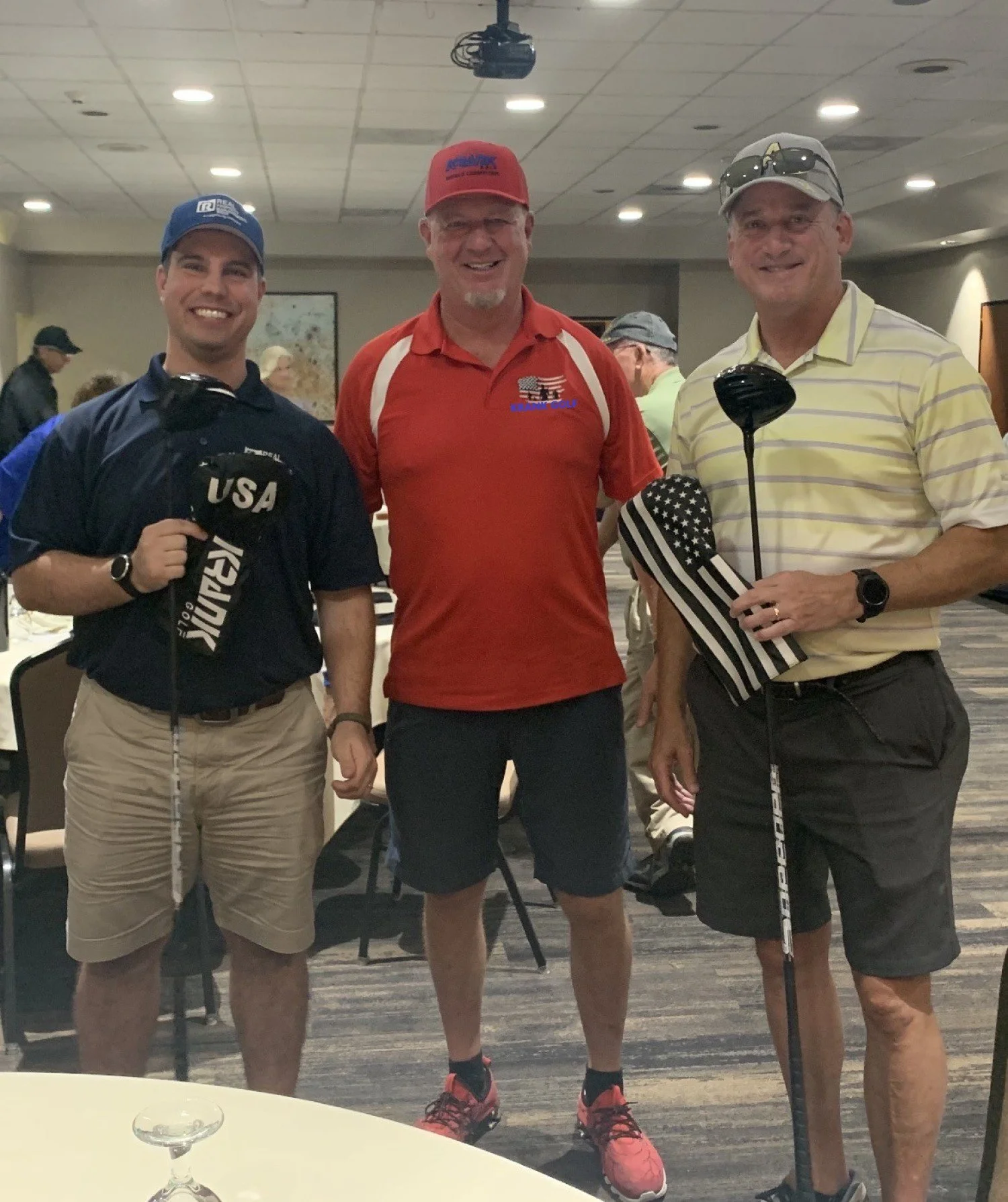 Three men standing together in a room, all holding golf clubs. One is wearing a black shirt with 'USA' and a cap, the second is in a red shirt with a golf-themed logo, and the third is in a yellow striped polo shirt with sunglasses on his cap. They are smiling and appear to be at a golf-themed event or gathering.