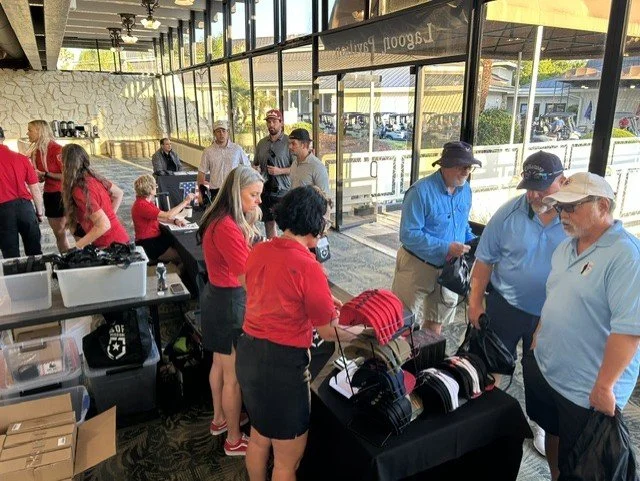 People shopping at a retail event inside a store with large glass windows, displaying hats, shirts, and other merchandise on tables.