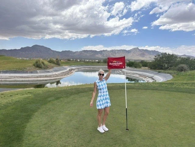A woman in a blue and white checkered dress holding a red flag on a golf course with a water hazard, mountains, and cloudy sky in the background.