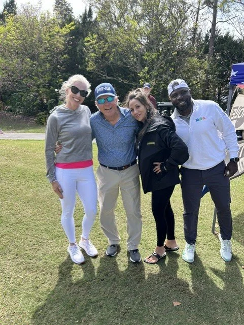 Four people standing outdoors on grass, smiling at the camera. Two women and two men, one of the women and the man wearing sunglasses, dressed casually. The background has trees and a tent with a Texas flag.